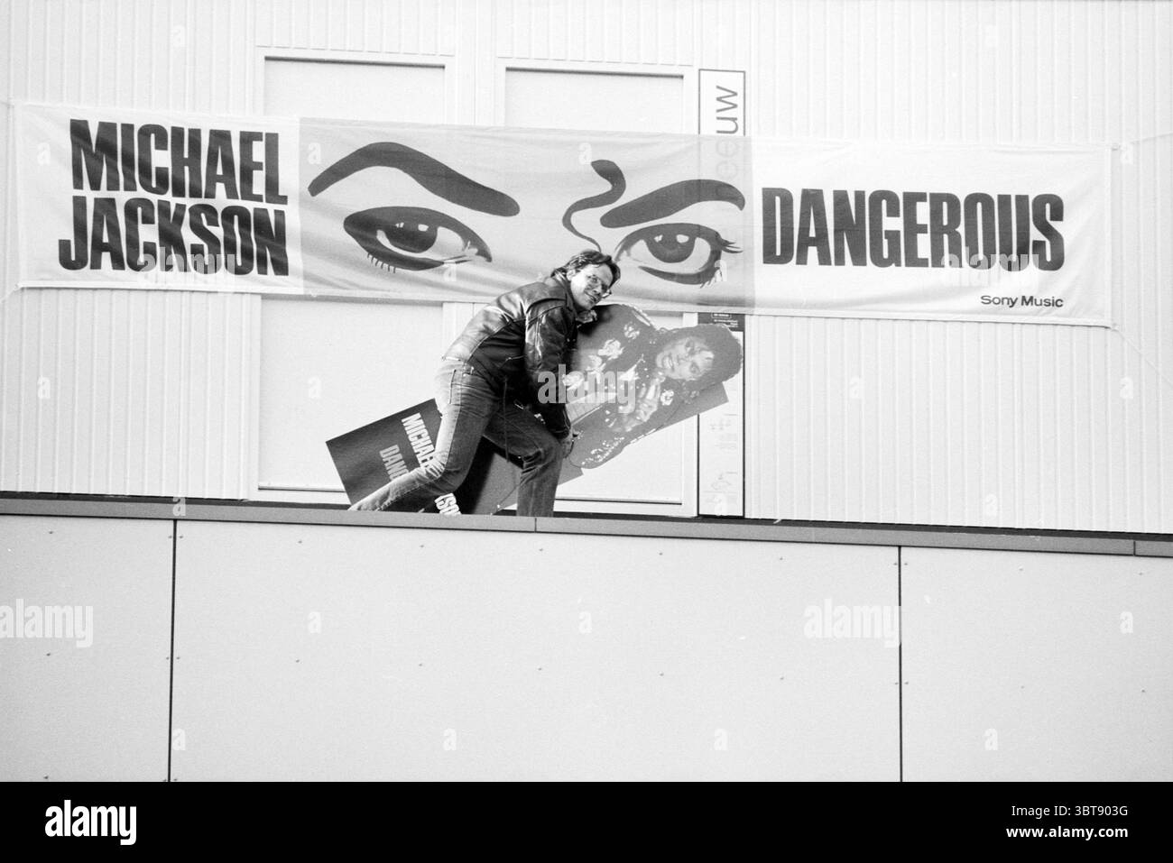 Gerard Rutte Sony Music, Whizgle News, Dutch Desk, pays-Bas, 1950 - 2000 le 19-11-1991. Voici les rubriques de l'image. Une scène saisissante se déroule, mettant en vedette une figure perchée sur le bord d'un toit. L’individu, vêtu d’une élégante veste en cuir noir, est intentionnellement engagé avec une grande pochette d’album qui semble incarner une présence musicale emblématique. La position de la personne suggère un moment de vénération ou d’admiration, contrastant avec la toile de fond vibrante d’une grande bannière aux yeux noirs et blancs évocateurs, qui attirent l’attention immédiate. Le cadre est urbain et un peu gritty, charac Banque D'Images