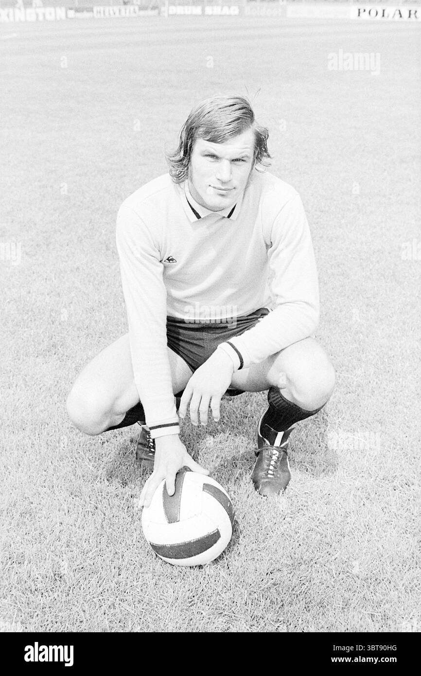 Portraitiste de football, Whizgle News, Dutch Desk, pays-Bas, 1950 - 2000 en 1972. L'image inclut ces rubriques. La scène montre un athlète masculin agenouillé sur un terrain herbeux, exsudant un comportement confiant et concentré. Il a des cheveux ondulés de longueur moyenne qui encadrent doucement son visage, et son expression est sérieuse mais accessible. Il porte un maillot à manches longues de couleur claire avec des rayures sombres aux poignets, jumelé à un short sombre qui souligne sa construction athlétique. Devant lui repose un ballon de football noir et blanc classique, poli et bien défini sur fond de vert éclatant Banque D'Images