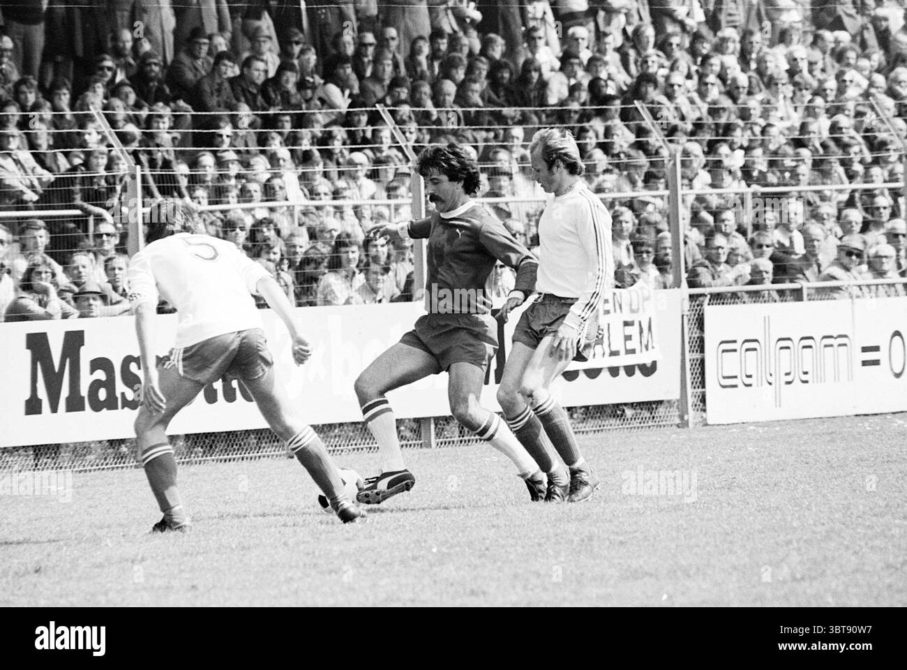 Haarlem - PSV Football Haarlem, Whizgle News, Dutch Desk, pays-Bas, 1950 - 2000 le 15-05-1977. L'image contient ces rubriques. La scène présente un moment dynamique sur un terrain de football, avec une énergie vibrante émanant des joueurs et de la foule. Au premier plan, deux athlètes sont pleinement engagés dans l’action. Un joueur, portant un maillot rouge vif avec des rayures blanches contrastées, semble manœuvrer habilement le ballon, tandis que le joueur adverse en blanc tente d'intercepter. L'herbe sous eux est un vert luxuriant, bien entretenu et fraîchement coupé, offrant une toile de fond vive pour Banque D'Images