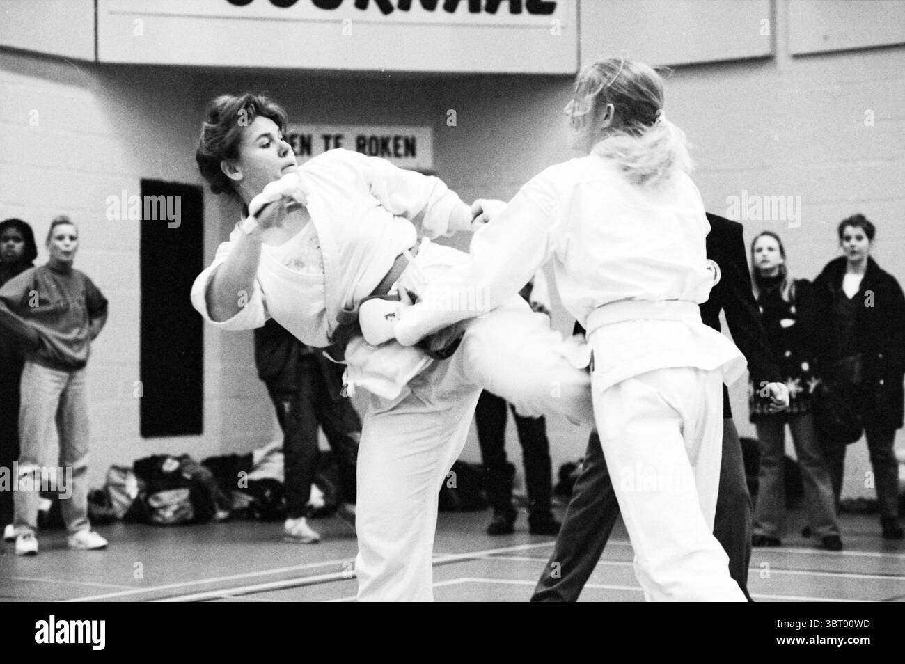 N.K. Karate Beverwijk Beverwijk pays-Bas, Whizgle News, Dutch Desk, pays-Bas, 1950 - 2000 le 27-02-1993. Voici les rubriques de l'image. Dans un cadre intérieur, une scène dynamique se déroule avec deux individus engagés dans des arts martiaux Les combattants portent des uniformes d'arts martiaux traditionnels, soulignant la nature formelle et disciplinée du sport. Un individu, exécutant un coup de pied élevé, montre un sentiment de puissance et de concentration, tandis que l'autre se tient sur la défensive, prêt à répondre. Autour d'eux, une collection de spectateurs peut être vue, certains engagés à regarder intentionnellement, tandis que d'autres app Banque D'Images
