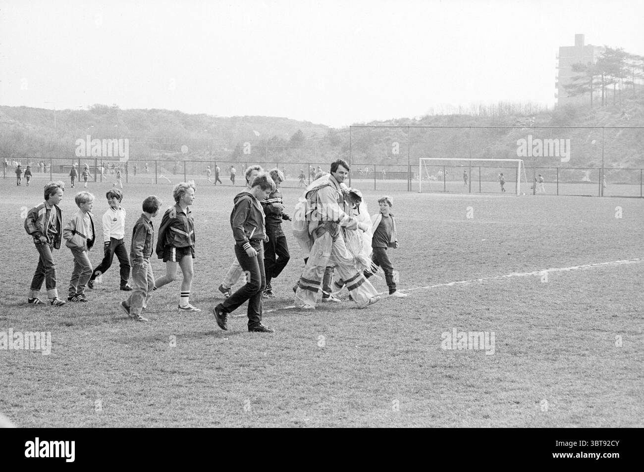 Velsen et ses environs parapente du jour de la Reine sur le terrain Stormvogels., Whizgle News, Dutch Desk, pays-Bas, 1950 - 2000. Ce sont les éléments de l'image. Dans un cadre extérieur animé, un groupe d'enfants et d'adultes se promène dans un champ herbeux, leurs marches marquant un chemin dans le gazon vert luxuriant. La scène est encadrée par des collines basses et vallonnées en arrière-plan, avec des contours doux d'arbres et d'arbustes, suggérant un paysage doux et naturel. La palette de couleurs prédominante est composée de différentes nuances de vert, reflétant l'herbe fraîchement tondue, aux côtés de gris et blancs frais du o Banque D'Images