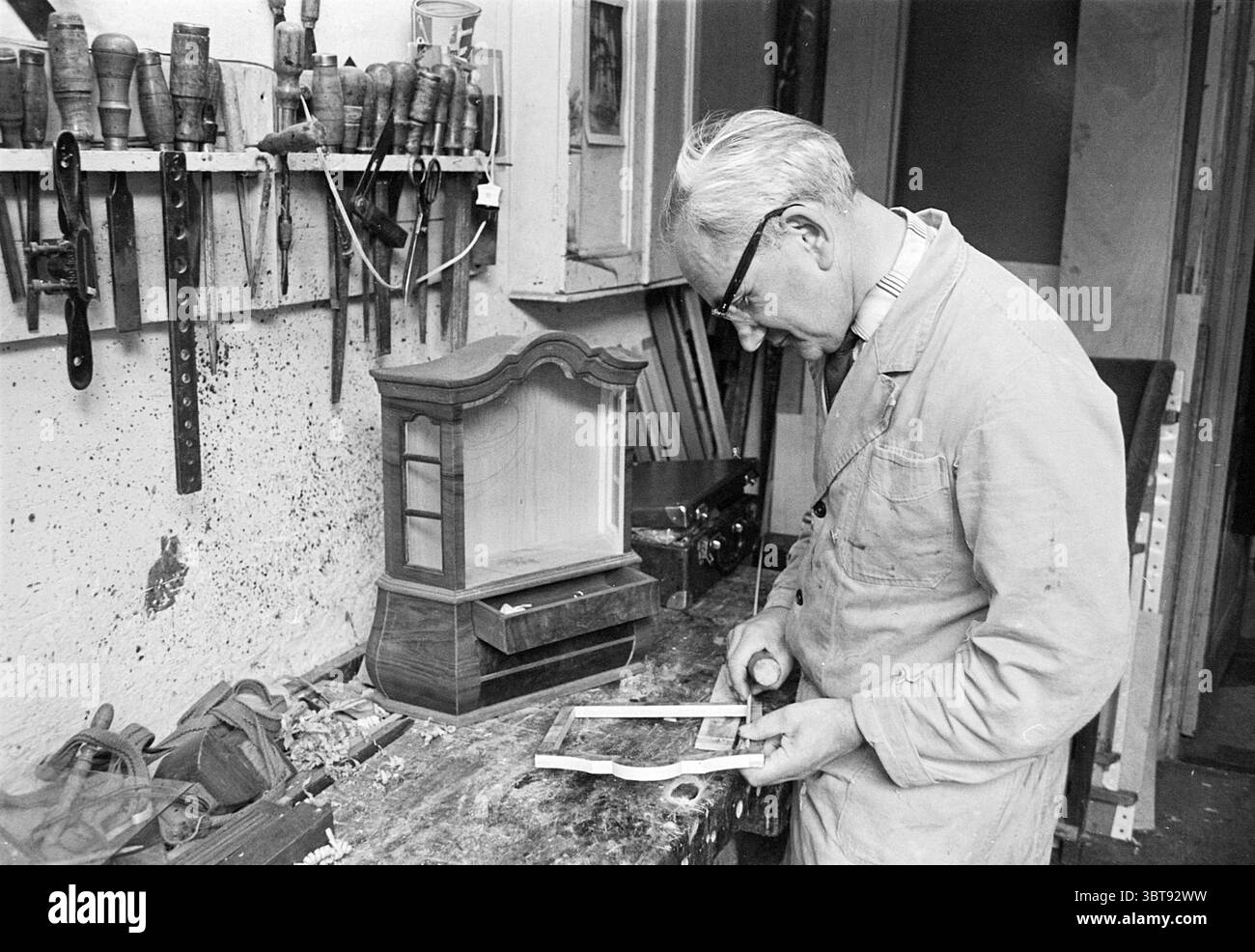 M. Jongbloed Persons, Whizgle News, Dutch Desk, pays-Bas, 1950 - 2000 le 05-07-1966. Voici les rubriques de l'image. Dans un atelier chaleureusement éclairé rempli d'un charme rustique, un artisan plus âgé, vêtu d'un manteau de couleur claire, est profondément concentré sur sa tâche. Ses cheveux argentés sont soigneusement peignés en arrière, et il porte des lunettes rondes qui reflètent la douce lueur de l'environnement. L'espace de travail est encombré mais organisé, affichant une gamme d'outils en bois accrochés au mur, leurs poignées portées depuis des années d'utilisation. Chaque outil est soigneusement placé, faisant allusion à l'expertise et à l'histoire de l'artisan. Banque D'Images