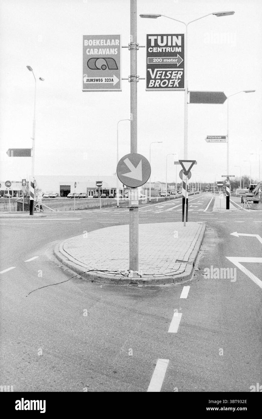 Panneaux publicitaires Velserbroek signe Velserbroek, Whizgle News, Dutch Desk, The Netherlands, 1950 - 2000 le 09-02-1990. Voici les rubriques de l'image. La scène présente un environnement principalement urbain caractérisé par des espaces ouverts et un réseau de routes. Au premier plan, un panneau de circulation circulaire avec une flèche indique un virage à gauche, soulignant le flux des véhicules. Ce signe est monté sur un poteau cylindrique, ce qui ajoute un point focal à la composition. Autour de l'intersection, plusieurs lampadaires hauts se dressent, leurs poteaux blancs créant des lignes verticales contre les couleurs plus subtiles o Banque D'Images