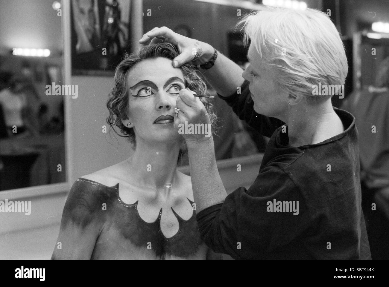 New look Body Painting Haarlem pays-Bas, Whizgle News, Dutch Desk, pays-Bas, 1950 - 2000 sur 30-08-1996. L'image contient ces rubriques. Dans un dressing légèrement éclairé, une scène saisissante se déroule. Une femme aux cheveux bouclés est assise à une vanité, son visage partiellement transformé avec un maquillage dramatique. La maquilleuse, avec des cheveux courts et clairs, se penche étroitement, appliquant des touches de finition à son maquillage des yeux. Les traits de la femme sont accentués par un eye-liner graphique audacieux qui s'étend vers l'extérieur, créant un effet théâtral. Sa peau est ornée de peinture corporelle complexe qui imite f Banque D'Images