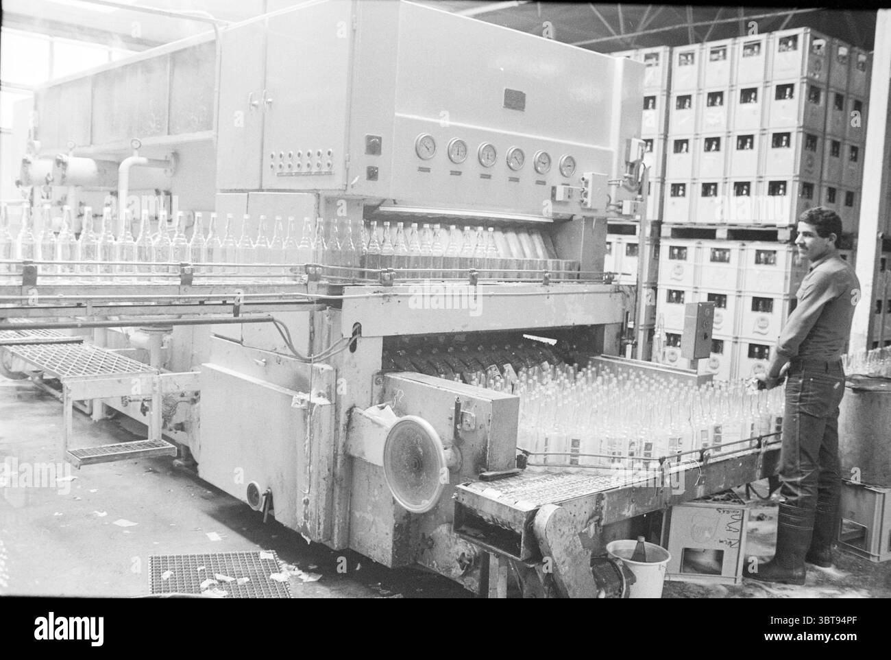 Lave-bouteilles Loots Lemonade Drinks and Liquor Stores, Whizgle News, Dutch Desk, pays-Bas, 1950 - 2000 le 21-06-1982. Voici les rubriques de l'image. La scène représente un environnement industriel, caractérisé par une grande configuration de machines conçues pour le traitement des bouteilles en verre. La caractéristique dominante est une machine importante, qui semble être une ligne d'embouteillage, avec de nombreuses bouteilles en verre transparent disposées symétriquement le long de sa bande transporteuse. La machine a une surface métallique et une série de cadrans de commande ou de boutons, faisant allusion à sa fonctionnalité complexe. Sur le côté gauche du Banque D'Images