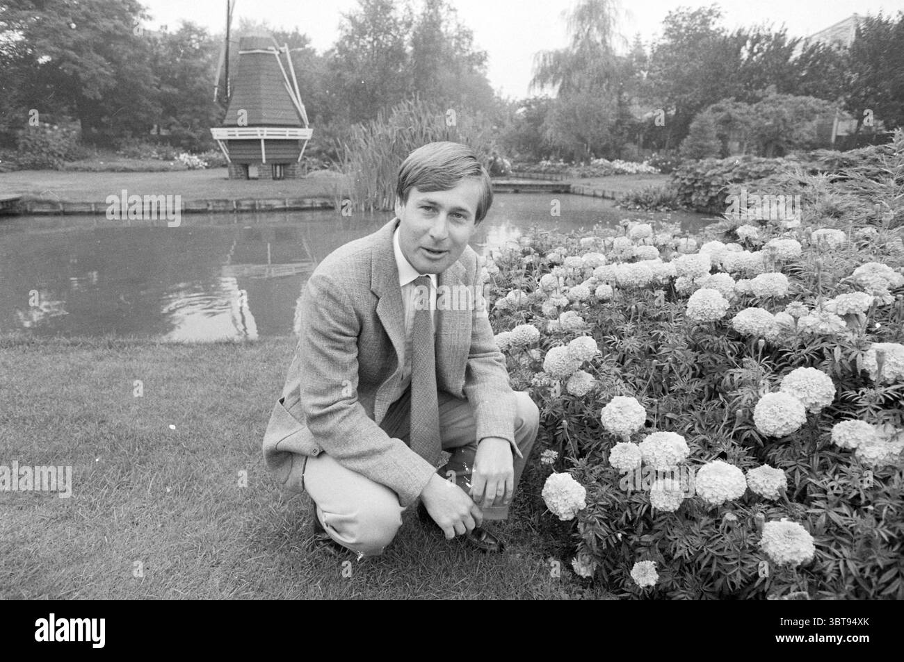 M. Leo Roozen dans le jardin Vogelenzang jardins et parcs Vogelenzang, Whizgle News, Dutch Desk, pays-Bas, 1950 - 2000 le 31-08-1983. Voici les rubriques de l'image. Une scène de jardin sereine se déroule, caractérisée par une étendue luxuriante de verdure méticuleusement entretenue. Au premier plan, un homme, vêtu d'un costume gris clair avec un blazer subtilement texturé, s'agenouille à côté d'une vivacité de fleurs. Les fleurs sont un mélange de couleurs douces – blancs et jaunes pâles – qui se démarquent du vert riche du feuillage et de l’herbe. Le fond révèle un étang tranquille, sa surface reflétant th Banque D'Images