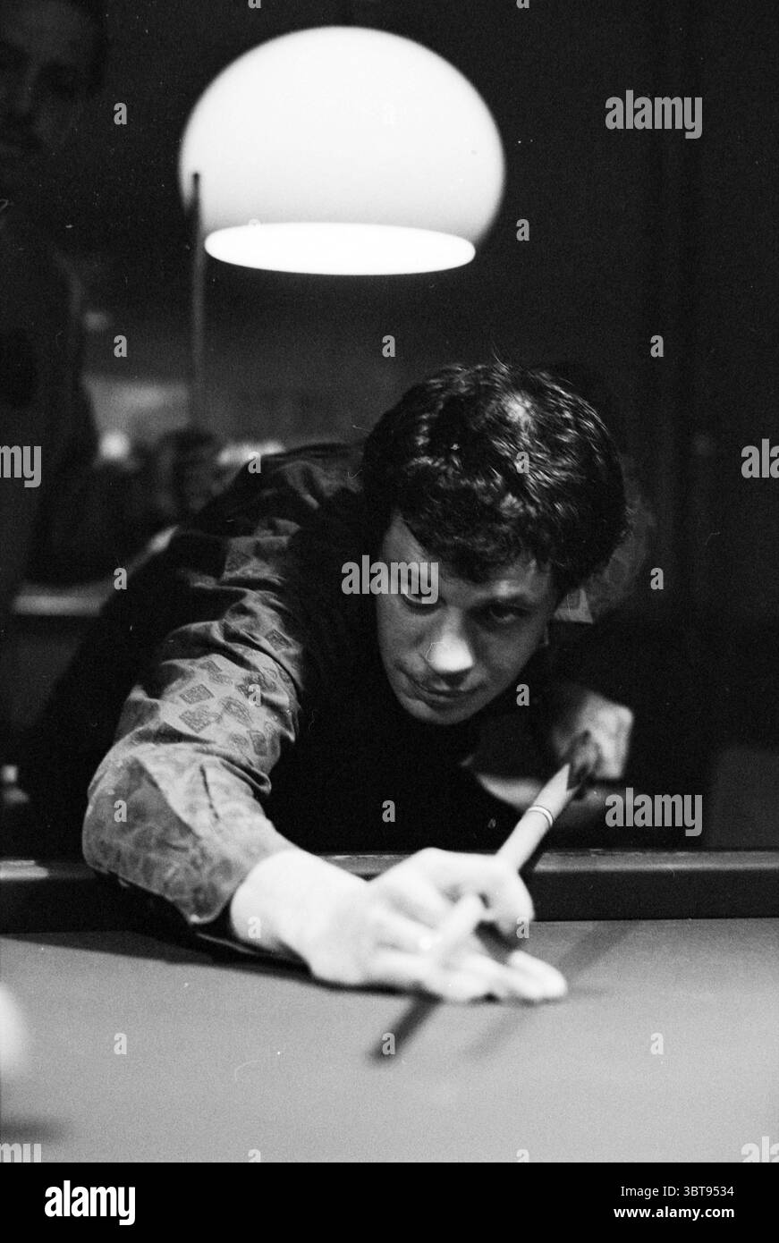 Disabled billard J. v. Beem, Whizgle News, Dutch Desk, pays-Bas, 1950 - 2000 on 13-03-1993. L'image inclut ces rubriques. Dans un cadre faiblement éclairé, l'accent est mis sur un jeune homme se concentrant intensément sur une partie de billard. Il se penche en avant au bord d’une table en feutre vert, sa posture suggérant un mélange de détermination et de concentration. La tenue de l'homme comporte une chemise à motifs avec des tons discrets, ajoutant à l'ambiance rétro de la scène. La table présente plusieurs boules de billard dans un tableau classique, vibrant sur la surface vert profond, avec un mélange de rouges, jaunes et bleus standi Banque D'Images