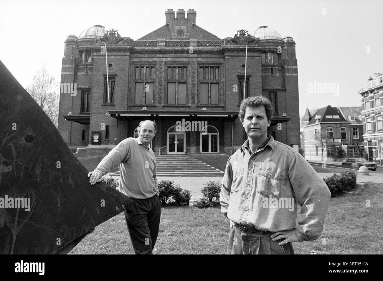 Messieurs Graaf et Monts devant le Schouwburg, Whizgle News, Dutch Desk, pays-Bas, 1950 - 2000 le 09-05-1991. Ces rubriques apparaissent dans l'image. Au premier plan, deux hommes se tiennent debout avec confiance, chacun portant des vêtements décontractés et légèrement décontractés. Un homme se penche avec désinvolture contre une grande sculpture en métal angulaire qui ajoute un contraste contemporain à l'architecture traditionnelle derrière eux. L'autre se tient debout, regardant directement le spectateur avec une expression sérieuse. Le fond est dominé par un grand bâtiment historique caractérisé par sa façade en briques ornées et son grand vent voûté Banque D'Images Messieurs Graaf et Monts devant le Schouwburg, Whizgle News, Dutch Desk, pays-Bas, 1950 - 2000 le 09-05-1991. Ces rubriques apparaissent dans l'image. Au premier plan, deux hommes se tiennent debout avec confiance, chacun portant des vêtements décontractés et légèrement décontractés. Un homme se penche avec désinvolture contre une grande sculpture en métal angulaire qui ajoute un contraste contemporain à l'architecture traditionnelle derrière eux. L'autre se tient debout, regardant directement le spectateur avec une expression sérieuse. Le fond est dominé par un grand bâtiment historique caractérisé par sa façade en briques ornées et son grand vent voûté Banque D'Images