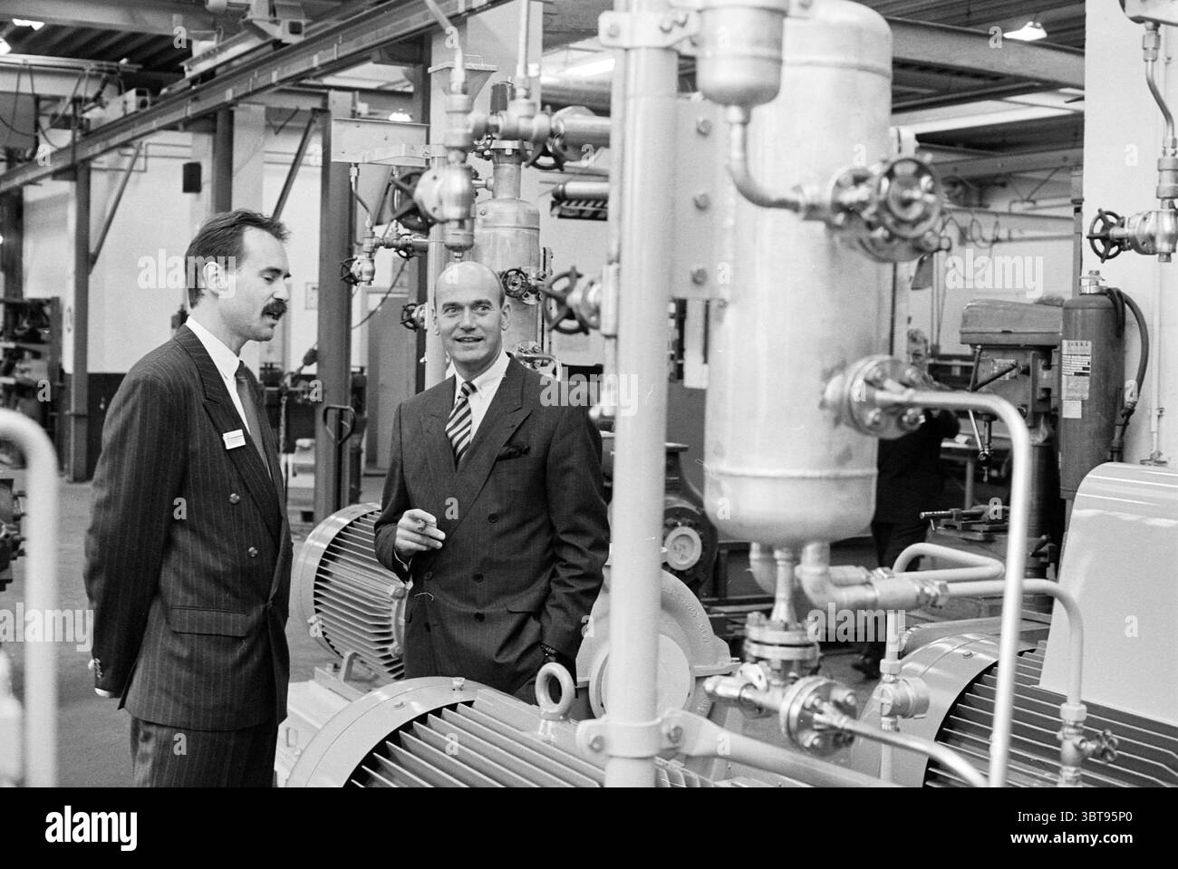 Ouverture de l'usine de pompes Zwanenburg, Whizgle News, Dutch Desk, pays-Bas, 1950 - 2000 le 13-04-1994. Ce sont les éléments de l'image. La scène capture un environnement industriel animé, caractérisé par son intérieur spacieux rempli de machines et d'équipements. L'environnement est principalement composé de structures métalliques, ce qui lui confère une sensation robuste et mécanique. Deux hommes se tiennent près de gros moteurs, engageant la conversation. Un homme est vêtu d'un costume rayé, exsudant un comportement professionnel. Son expression exprime de l'intérêt lorsqu'il écoute attentivement. L'autre homme, également formellement vêtu Banque D'Images