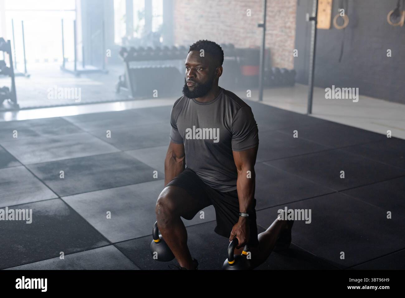 Homme afro-américain portant des vêtements de sport agenouillant sur un genou et soulevant des kettlebells dans la salle de gym Banque D'Images