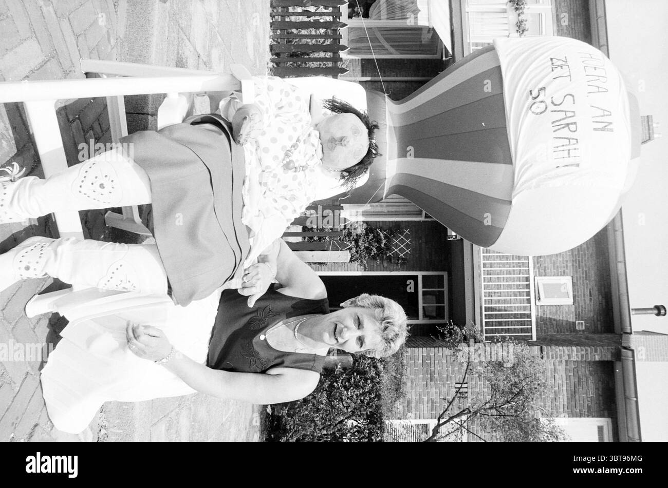 MSRS an v. GE@le@ avec Sara IJm. [Van Gevle] IJmuiden pays-Bas, Whizgle News, Dutch Desk, pays-Bas, 1950 - 2000 le 08-06-1992. L'image inclut ces rubriques. Dans un cadre extérieur lumineux, une scène joyeuse se déroule. Une femme, avec des cheveux de couleur claire coiffés soigneusement, est assise sur un chemin de pierre. Elle porte une blouse légère et a une expression chaleureuse et accueillante. À côté d'elle se trouve un personnage de ballon ludique et surdimensionné ressemblant à une figure caricaturale. Ce personnage a un visage arrondi et joyeux et est habillé dans une tenue colorée, avec des rayures rouges et blanches proéminentes sur sa tenue th Banque D'Images