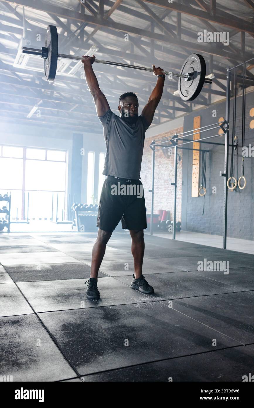 Homme afro-américain soulevant la barre chargée au-dessus de la tête dans les vêtements athlétiques à la salle de gym avec des anneaux Banque D'Images