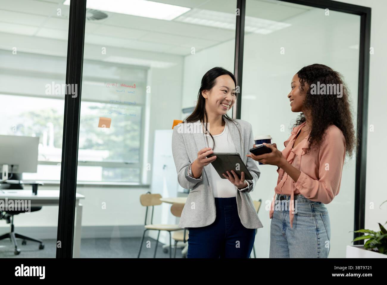 Diverses collègues féminines debout près de la cloison dans le bureau avec tablette et tasse à café discutant du travail Banque D'Images