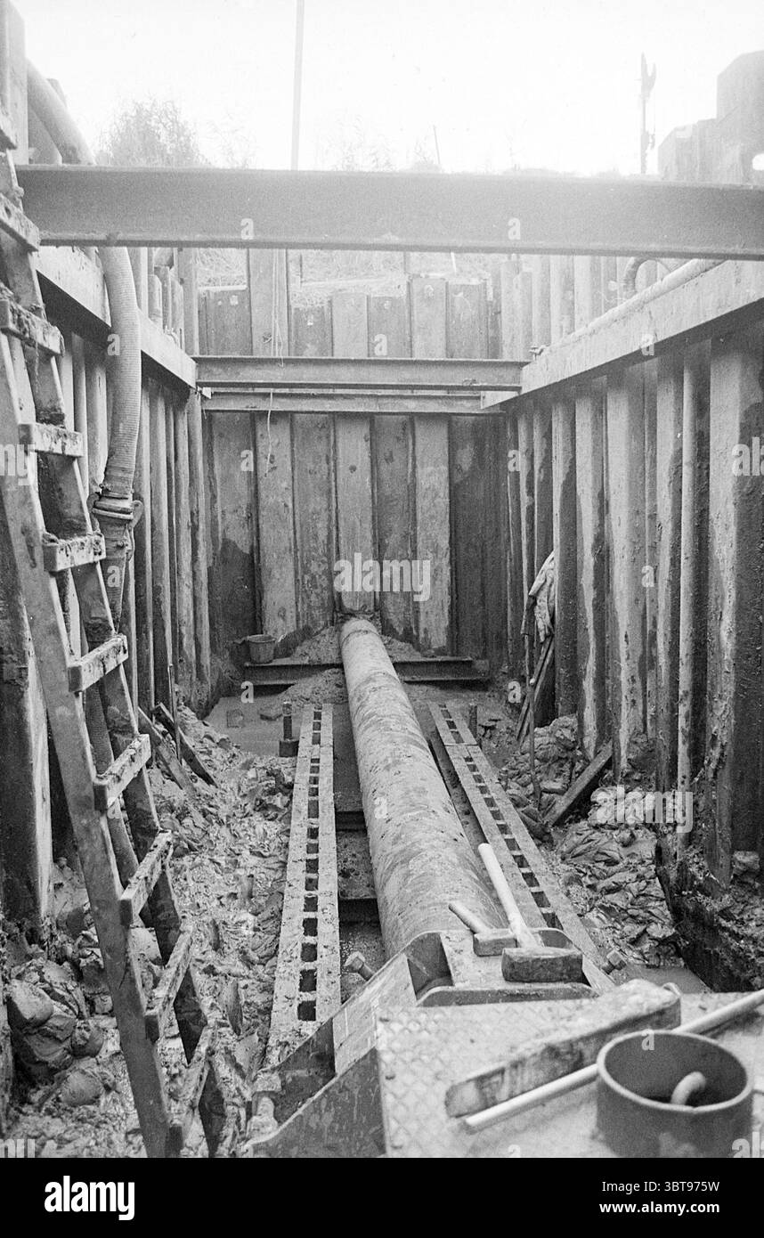 Remblai ferroviaire de pressage de tuyaux Enkhuizen de Ruiter B.V. Halfway, Whizgle News, Dutch Desk, pays-Bas, 1950 - 2000 on 24-10-1973. Ces rubriques apparaissent dans l'image. La scène représente un chantier de construction souterrain, caractérisé par une tranchée rectangulaire entourée de supports et de murs en bois. L'éclairage est assez faible, suggérant soit tôt le matin ou en fin d'après-midi, avec une lumière douce et diffuse éclairant l'espace. Au centre, un long tuyau cylindrique s'étend le long de la base de la tranchée, sa texture rugueuse et industrielle, contrastant avec les matériaux de terre environnants. Le Banque D'Images