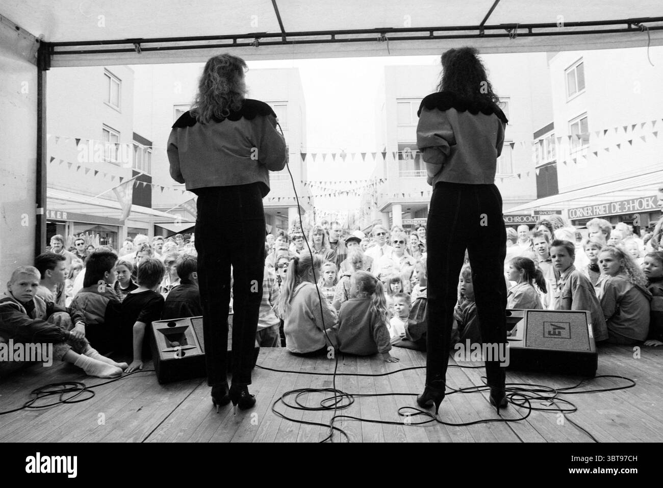 Maywood Velserbroek, Whizgle News, Dutch Desk, pays-Bas, 1950 - 2000 sur 28-06-1991. Voici les rubriques de l'image. Dans un cadre extérieur animé, une petite scène est encadrée par des bâtiments de couleur claire avec de grandes fenêtres, suggérant un environnement urbain animé d'activité. Deux interprètes se tiennent dos au spectateur, tous deux vêtus de tenues noires élégantes ornées d'épaulettes frappantes qui ajoutent une sensation de drame et de flair. Leurs cheveux sont coiffés dans des boucles lâches, contribuant à l'aspect dynamique global. La foule devant eux est diverse ; les gens de différents âges sont captivés Banque D'Images