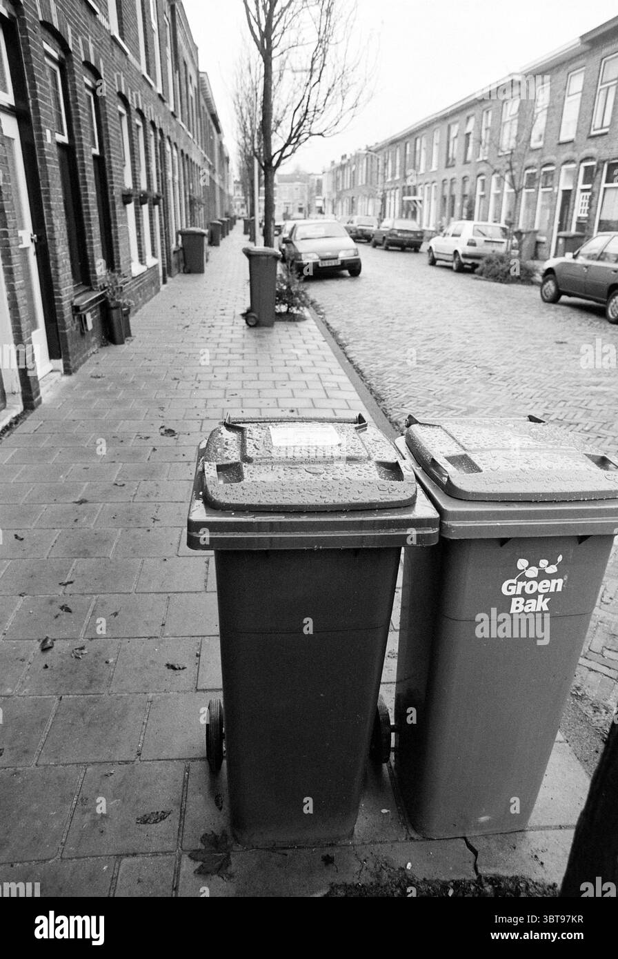 Seaux roulants dans la rue Bankastraat Haarlem Bankastraat pays-Bas, Whizgle News, Dutch Desk, pays-Bas, 1950 - 2000 on 01-12-1993. L'image contient ces rubriques. La scène représente une rue urbaine calme bordée de modestes bâtiments en briques. Le trottoir est mouillé, laissant entendre une pluie récente, et il brille sous une lumière douce et silencieuse qui suggère un ciel couvert. Au premier plan, deux poubelles à roulettes sont positionnées côte à côte, leurs couleurs contrastant avec l'environnement en niveaux de gris : elles semblent être d'un vert foncé et d'une teinte plus claire. Les bacs sont situés sur le côté gauche du côté Banque D'Images