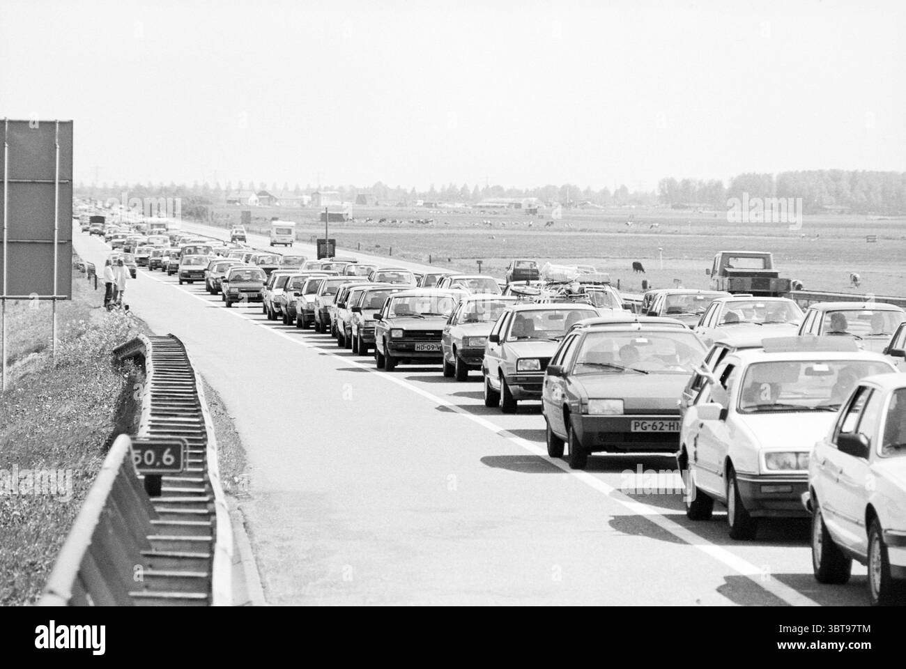 Embouteillages à Velsertunnel et ferry occupé IJmuiden occupé Velsertunnel IJmuiden Rijksweg A22 pays-Bas, Whizgle News, Dutch Desk, pays-Bas, 1950 - 2000 on 24-05-1987. Ces rubriques sont illustrées dans l'image. La scène représente une longue file de véhicules, étroitement emballés ensemble, s'étendant à travers une autoroute. Les voitures sont principalement des modèles vintage, présentant une variété de formes et de tailles, avec une palette de couleurs monotone qui met en valeur leurs caractéristiques dans des tons de gris. La route est flanquée d'une médiane herbeuse sur un côté, ajoutant une touche de texture naturelle contre l'étendue asphaltée. Dans le Banque D'Images