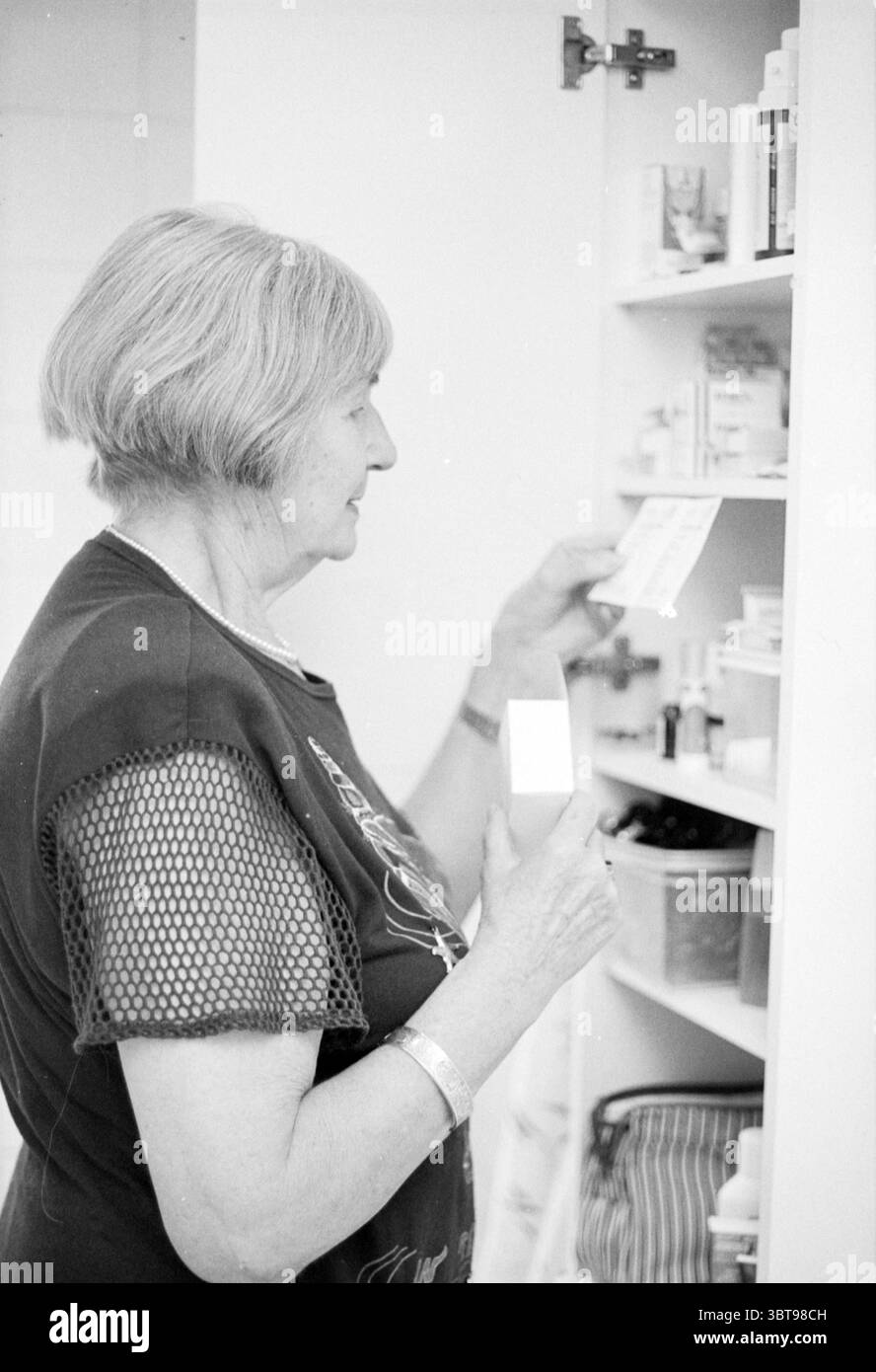Older + médicaments, Whizgle News, Dutch Desk, pays-Bas, 1950 - 2000 le 24-08-1995. L'image contient ces rubriques. Dans une salle de bain éclairée, une femme âgée se tient à côté d'une armoire blanche remplie de bouteilles et de contenants divers. Le cabinet est ouvert, révélant une gamme de produits de soins personnels qui suggèrent une routine quotidienne. La femme a une coiffure argentée qui encadre son visage, et elle porte un haut sombre avec une texture en maille, contrastant avec la sensation globale de clarté et de propreté de l'espace. Son expression semble concentrée lorsqu'elle examine une petite carte, en vérifiant peut-être th Banque D'Images