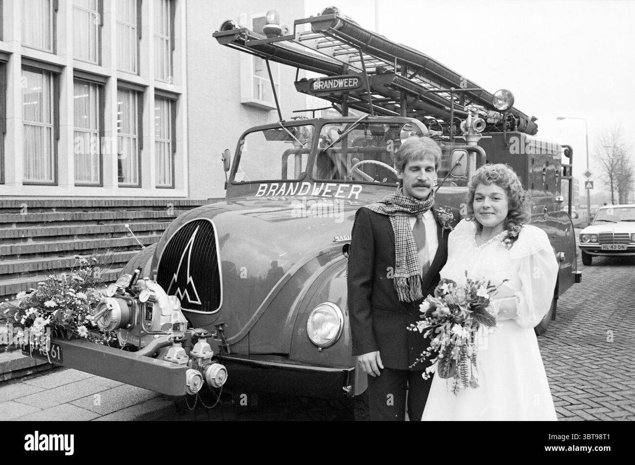 Mariage de Mr. Michel avec le vieux pompier IJm. Mariage IJmuiden pays-Bas, Whizgle News, Dutch Desk, pays-Bas, 1950 - 2000 le 16-12-1983. L'image montre ces rubriques. La scène capture un moment de célébration devant un camion de pompiers vintage. Le cadre est urbain, probablement à l'extérieur d'un bâtiment municipal, avec une toile de fond texturée de formations carrées en pierre et de grandes fenêtres rectangulaires. Au premier plan, un couple se tient côte à côte, exsudant joie et excitation. Le marié, vêtu d'un costume sombre avec une chemise de couleur claire et une cravate frappante, a une coiffure classique qui ajoute à la Banque D'Images