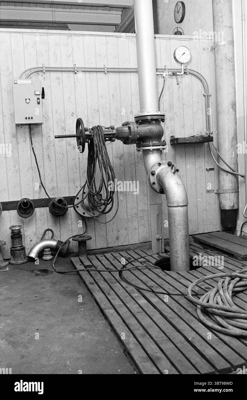 comité d'entreprise et 2 valves de Ruiter B.V. Halfway, Whizgle News, Dutch Desk, pays-Bas, 1950 - 2000 on 11-12-1973. L'image montre ces rubriques. La scène capture un cadre industriel fonctionnel, dominé par un mélange d'éléments métalliques et de bois. Un grand tuyau en acier s'étend bien en évidence à partir du sol, sa surface montrant des signes d'usure avec un lustre légèrement terne. Le tuyau mène à un mécanisme de soupape, qui est angulaire et robuste, indiquant qu'il joue un rôle critique dans le contrôle de l'écoulement du fluide. À proximité, une longueur de tuyau noir enroulé est soigneusement enroulée autour d'une bobine circulaire, suggérant rea Banque D'Images
