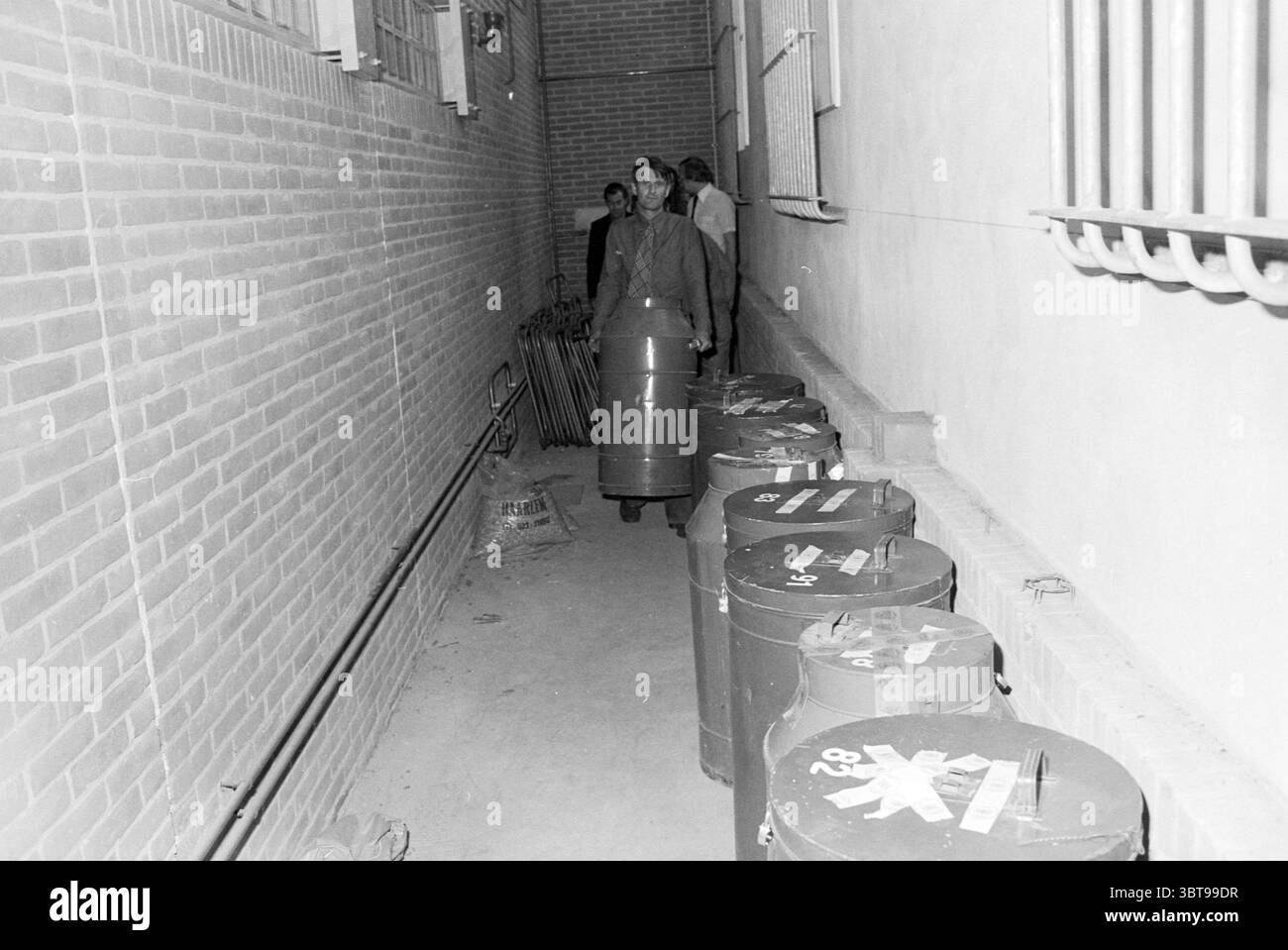 Urnes de vote stockées au commissariat de police de Haarlem, Whizgle News, Dutch Desk, pays-Bas, 1950 - 2000 le 07-06-1979. Ce sont les éléments de l'image. La scène se déroule dans un couloir étroit et faiblement éclairé caractérisé par son atmosphère industrielle. Les murs sont construits à partir de briques rouges rugueuses, prêtant une texture granuleuse à l'environnement. Le sol est en béton, et l'espace semble confiné, ce qui souligne un sentiment d'isolement. Le long d'un côté du couloir, une série de grands conteneurs cylindriques sont alignés. Leurs surfaces sont métalliques, peintes dans des couleurs sombres comme le vert ou le gris, Banque D'Images Urnes de vote stockées au commissariat de police de Haarlem, Whizgle News, Dutch Desk, pays-Bas, 1950 - 2000 le 07-06-1979. Ce sont les éléments de l'image. La scène se déroule dans un couloir étroit et faiblement éclairé caractérisé par son atmosphère industrielle. Les murs sont construits à partir de briques rouges rugueuses, prêtant une texture granuleuse à l'environnement. Le sol est en béton, et l'espace semble confiné, ce qui souligne un sentiment d'isolement. Le long d'un côté du couloir, une série de grands conteneurs cylindriques sont alignés. Leurs surfaces sont métalliques, peintes dans des couleurs sombres comme le vert ou le gris, Banque D'Images