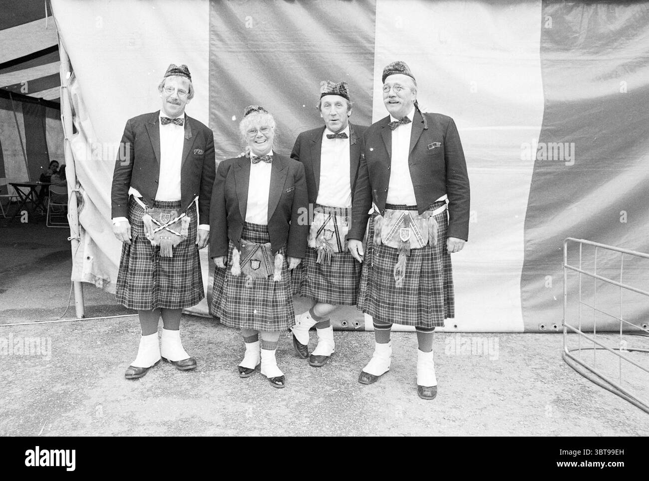 4 clowns Circus Mullens Rotterdam Circus, Whizgle News, Dutch Desk, pays-Bas, 1950 - 2000 on 10-05-1988. L'image contient ces rubriques. Dans un cadre extérieur animé, quatre personnes se tiennent ensemble, chacune présentant une tenue écossaise traditionnelle. Leurs tenues se composent de kilts à longueur de genou ornés d'un motif écossais audacieux dans des teintes de vert, bleu et noir, ce qui ajoute une texture riche à la scène. Les hommes portent des vestes sur mesure, complétées par des chemises blanches et des noeuds papillon, tandis que la femme partage un style similaire, son kilt associé à une veste ajustée, lui donnant un clin d'œil à l'ensemb classique Banque D'Images