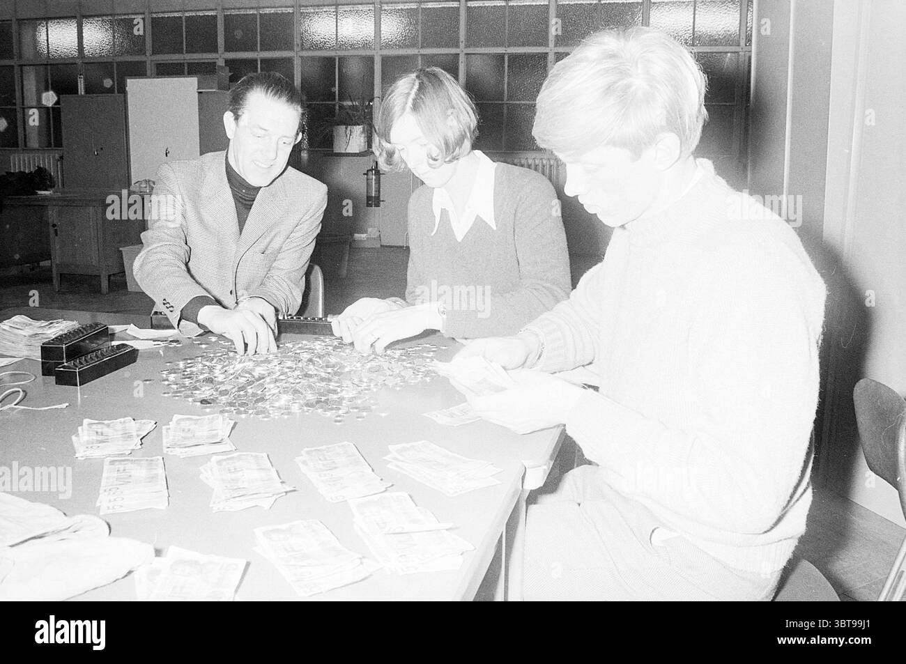 Carnival concert Hall Carnival, Whizgle News, Dutch Desk, pays-Bas, 1950 - 2000 le 13-02-1971. L'image contient ces rubriques. Dans une pièce faiblement éclairée, trois individus sont rassemblés autour d'une grande table encombrée d'un assortiment de pièces de monnaie et de piles de papier-monnaie. L'atmosphère semble concentrée et engagée, suggérant qu'ils sont impliqués dans le comptage ou le tri de l'argent. La première personne à gauche, un homme vêtu d'un blazer à motifs, se penche étroitement, examinant attentivement les pièces devant lui. Ses cheveux sont coiffés soigneusement, et il semble absorbé dans la tâche, mettant en valeur un look de c Banque D'Images