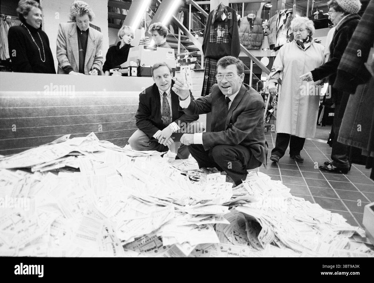 Tirage au sort au magasin de vêtements de Lords Heemstede Miscellaneous Lottery Heemstede pays-Bas, Whizgle News, Dutch Desk, pays-Bas, 1950 - 2000 on 14-11-1988. L'image contient ces rubriques. Dans un environnement de vente au détail animé, une scène encombrée se déroule sur un sol carrelé où une grosse pile de papier, par exemple des reçus ou du matériel promotionnel, se déverse, créant un contraste frappant avec les carreaux gris polis. Deux hommes s'agenouillent à côté du tas, l'un faisant des gestes animés avec un sourire tandis que l'autre regarde avec une expression perplexe, tous deux vêtus de costumes qui suggèrent un décontracté mais professionnel à Banque D'Images