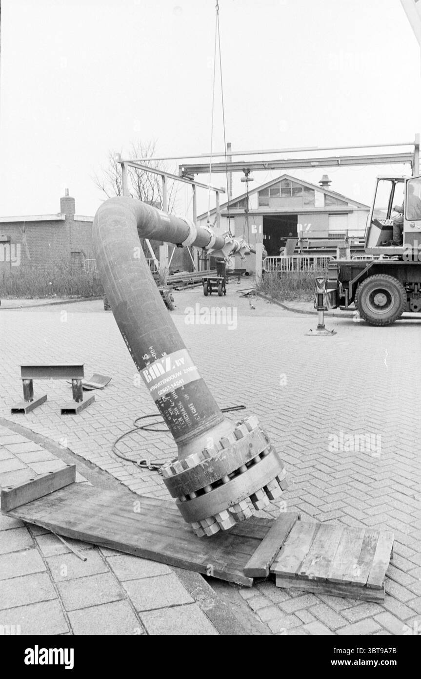 Tuyau de 36 mètres de long pour plate-forme de forage BMZ pose des tuyaux, Whizgle News, Dutch Desk, pays-Bas, 1950 - 2000 sur 06-04-1984. L'image contient ces rubriques. Dans un cadre industriel côtier, un grand tuyau métallique domine la scène. Le tuyau est courbé et semble lourd, reposant sur une plate-forme en bois qui supporte son poids. Il a une surface texturée, mettant en valeur l'usure de l'utilisation précédente, et est peint dans une couleur sourde, probablement gris ou métal rouillé. À proximité, une grue se dresse haute, partiellement englouti dans le fond, indiquant des opérations de levage actives. L'atmosphère générale est animée mais Banque D'Images