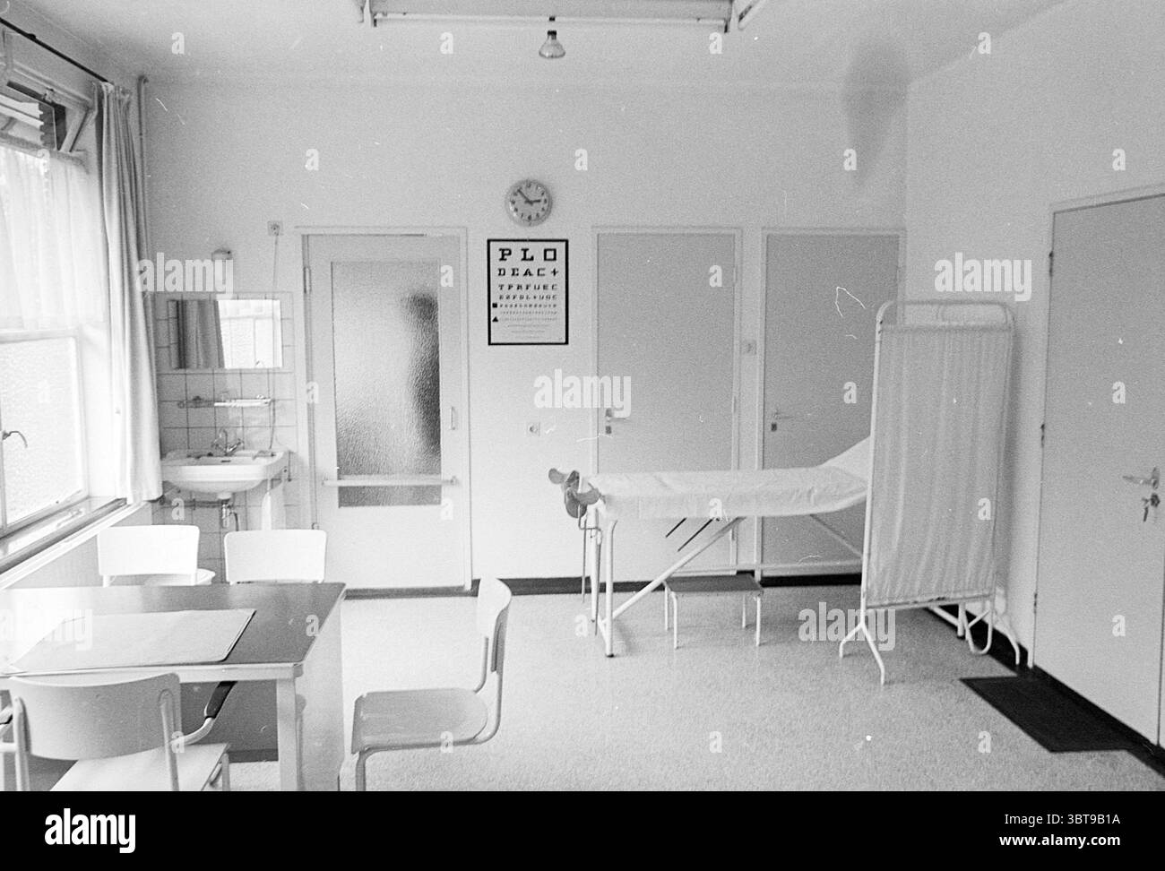 Salle de consultation Health House Heemstede Lieven de Keylaan pays-Bas, Whizgle News, Dutch Desk, pays-Bas, 1950 - 2000 on 13-08-1971. L'image inclut ces rubriques. La scène représente une salle clinique caractérisée par un design épuré et minimaliste. Les murs sont peints dans un blanc doux, contribuant à un sentiment de propreté et de stérilité. La lumière naturelle pénètre par une grande fenêtre partiellement couverte de rideaux transparents, projetant de douces ombres et créant une atmosphère calme. D'un côté, un petit évier est monté sur le mur, reflétant l'aspect pratique, tandis qu'une série de portes fermées, chacune Banque D'Images