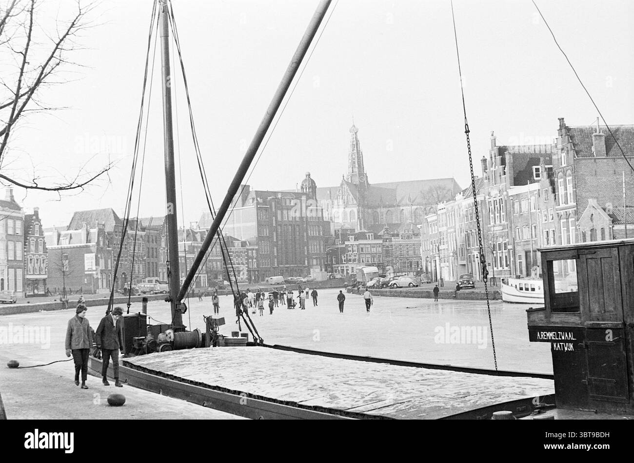 Patinage à Haarlem et Ice Club Nova Zembla à Spaarndam patinoire hockey sur glace, Whizgle News, Dutch Desk, pays-Bas, 1950 - 2000 on 26-12-1961. Ce sont les éléments de l'image. La scène représente une zone tranquille et hivernale de front de mer, caractérisée par une palette monochromatique de noirs, blancs et nuances de gris, évoquant une ambiance sereine et quelque peu nostalgique. Au premier plan, un grand bateau à coque sombre et à mât imposant domine le côté gauche, sa surface partiellement gelée, suggérant la présence de glace sur l’eau. À droite, un étroit sentier glacé mène à partir de l'eau Banque D'Images
