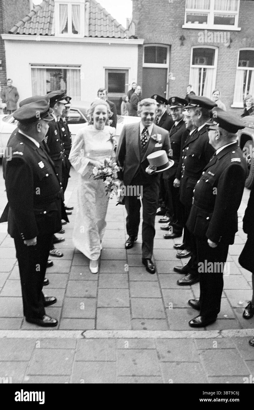Mariage des pompiers., Whizgle News, Dutch Desk, pays-Bas, 1950 - 2000. L'image contient ces rubriques. Dans une scène animée en plein air, un couple de jeunes mariés marche ensemble dans une rue pavée, rayonnant de joie et de célébration. La mariée, vêtue d'une robe blanche classique ornée de dentelle complexe, tient un bouquet de fleurs fraîches qui ajoute une touche de couleur à son ensemble. Ses cheveux sont élégamment coiffés, et elle porte un sourire radieux qui capture l'essence du moment. À côté d'elle, le marié apparaît dapper dans un costume pointu complété par un élégant chapeau de couleur claire, contribuant à un raffinement Banque D'Images