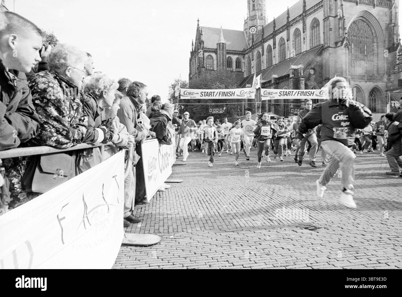Troop run Haarlem Grote Markt pays-Bas, Whizgle News, Dutch Desk, pays-Bas, 1950 - 2000 on 10-1992. L'image contient ces rubriques. La scène capture un moment énergique lors d'un événement de course à pied, dans un contexte historique avec une grande cathédrale, sa façade en pierre détaillée et ses flèches imposantes reflétant un sens de la tradition. Le premier plan est animé par une foule de spectateurs, leurs expressions étant un mélange d'excitation et d'anticipation alors qu'ils se penchent sur une barrière qui les sépare de la course. Plusieurs coureurs sont en mouvement, leurs corps inclinés vers l'avant, suggérant de la vitesse Banque D'Images