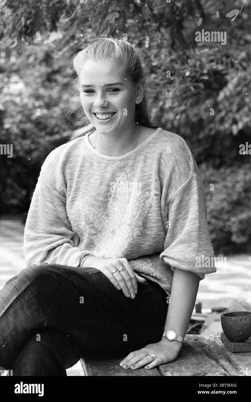 Leonie Voelink aspirante star de cinéma filmant des enregistrements de films, Whizgle News, Dutch Desk, pays-Bas, 1950 - 2000 le 07-09-1986. Voici les rubriques de l'image. Dans un cadre extérieur serein, une jeune femme s’assoit confortablement sur un banc en bois rustique. Elle a un sourire éclatant qui rayonne de chaleur et d'accessibilité. Son pull en tricot surdimensionné de couleur claire contraste doucement avec son Jean foncé, créant un look décontracté mais chic. Autour d'elle, un fond luxuriant de verdure ajoute une touche vibrante, avec diverses nuances de vert des feuilles et des branches qui l'encadrent. L'atmosphère se sent détendue et Banque D'Images