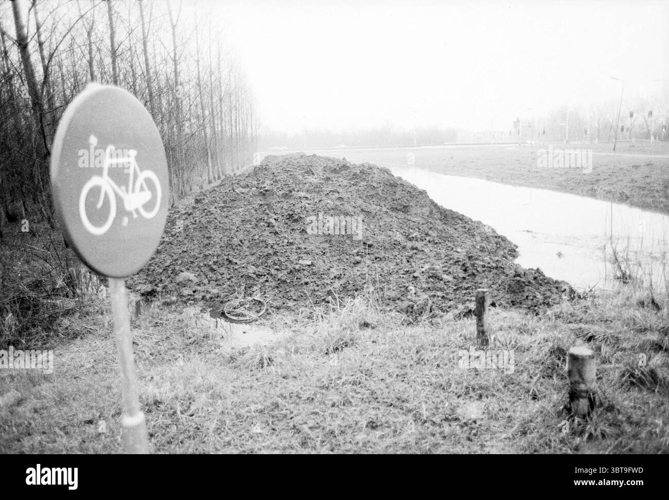 Espace de loisirs Spaarnwoude, Whizgle News, Dutch Desk, pays-Bas, 1950 - 2000 on 01-1988. L'image contient ces rubriques. La scène présente un paysage un peu désolé principalement dans les nuances de gris, lui donnant une atmosphère sombre et sombre. Un grand monticule de terre, probablement une excavation récente, domine le premier plan. Sa surface irrégulière et robuste contraste avec la douceur du sol environnant. Sur le côté gauche, un panneau indiquant une piste cyclable se dresse, sa forme circulaire mise en évidence sur la toile de fond de la butte de terre. L'icône de vélo simpliste du panneau ajoute une touche de colo Banque D'Images