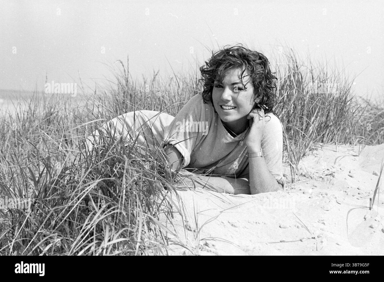 Simone Henning Miss Beach Pageant on the Beach Beach and Beaches Elections, Whizgle News, Dutch Desk, pays-Bas, 1950 - 2000 on 05-07-1983. L'image inclut ces rubriques. Dans cette scène, une femme est couchée sur une plage de sable, entourée de touffes d'herbe verte qui fournissent un cadre naturel. Ses cheveux foncés et bouclés sont émoussés, attrapant la douce brise, et elle arbore une tenue décontractée et de couleur claire qui contraste avec le sable environnant. Les couleurs présentes sont principalement atténuées, avec les tons doux du sable se mélangeant parfaitement aux teintes claires du ciel. L'atmosphère dégage un sen Banque D'Images