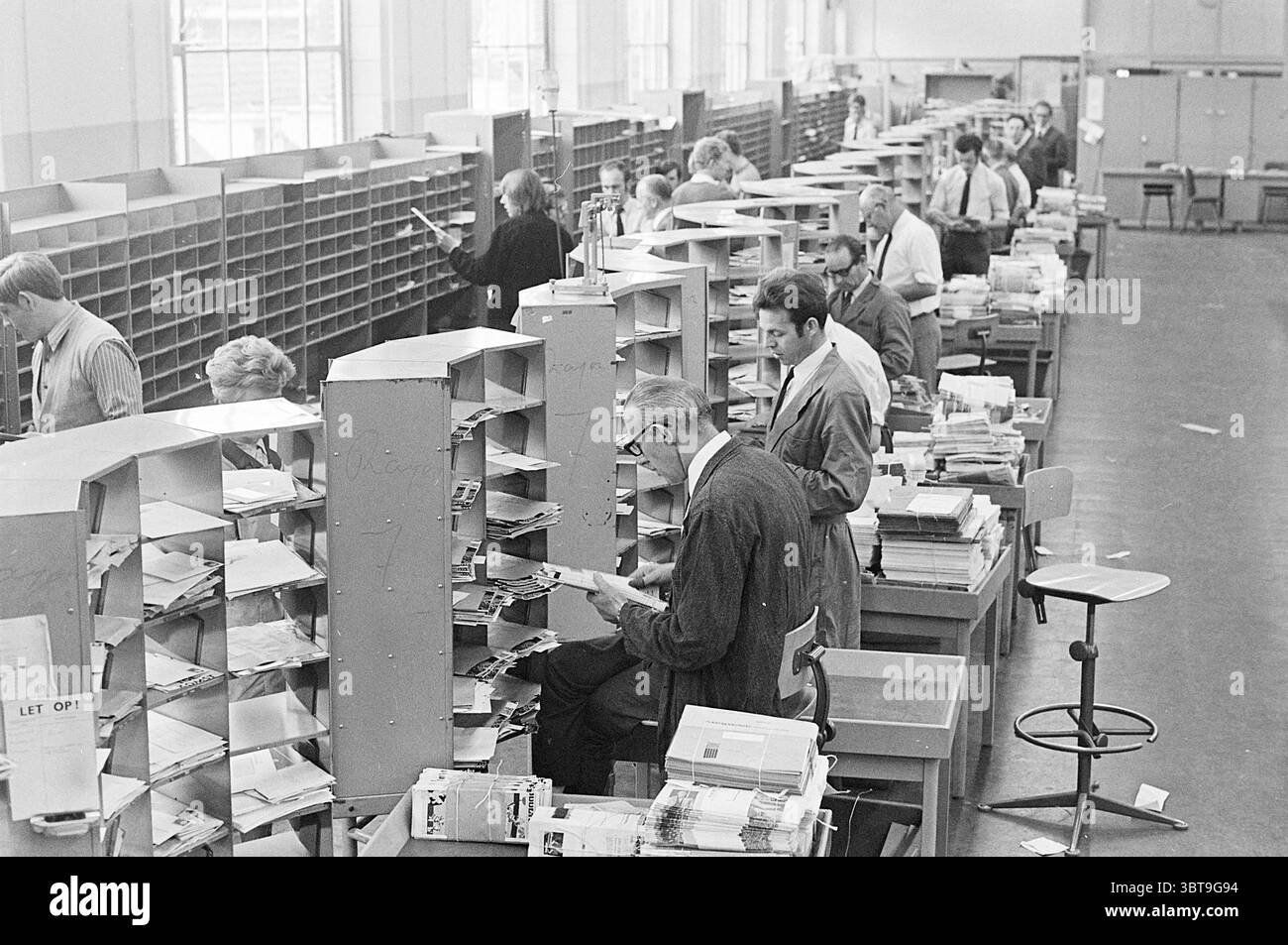 Tri PTT PTT postal services City mail, Whizgle News, Dutch Desk, pays-Bas, 1950 - 2000 on 15-06-1970. Ces rubriques apparaissent dans l'image. La scène représente un environnement intérieur occupé, probablement une installation postale ou de bureau, caractérisé par des rangées de bureaux de tri remplis de compartiments organisés. Diverses personnes sont engagées dans leurs tâches, entourées d'un sentiment d'activité ciblée. Les couleurs dominantes sont atténuées, avec des nuances de gris et de beige, suggérant un cadre utilitaire. Les murs sont de couleur claire, contribuant à une atmosphère lumineuse mais quelque peu stérile. Les grandes fenêtres permettent n Banque D'Images