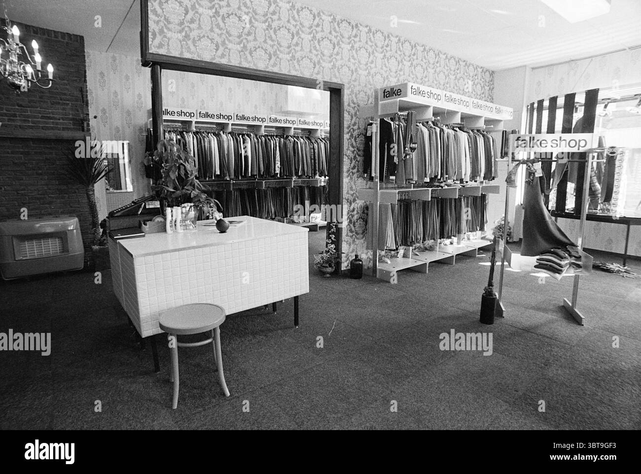 Intérieur Falke Shop Interior, Whizgle News, Dutch Desk, pays-Bas, 1950 - 2000 sur 29-08-1972. L'image contient ces rubriques. La scène représente un espace de vente au détail confortable, principalement axé sur les textiles et les vêtements. Les murs sont ornés d'un design de papier peint vintage, créant une atmosphère douce et accueillante qui contraste avec l'obscurité des produits exposés. Sur le côté gauche, un grand écran présente des rouleaux de tissu soigneusement disposés dans différentes couleurs et motifs, mettant en valeur un spectre allant des tons de terre atténués aux teintes vibrantes. L'éclairage dans l'espace est doux et diffus, peut-être à venir fr Banque D'Images