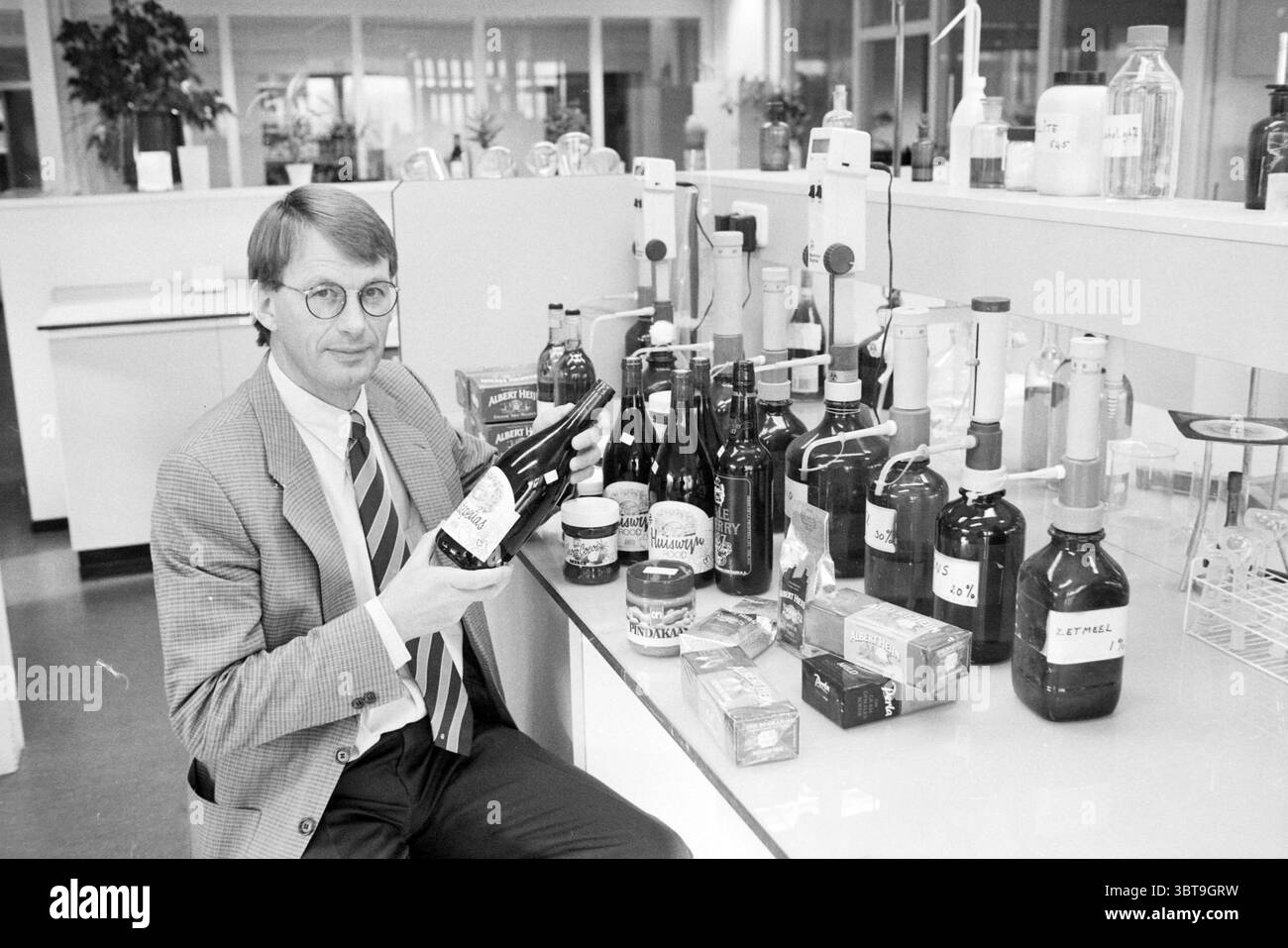 Mr. Oosterhuis Marvelo Zaandam Zaandam, Whizgle News, Dutch Desk, pays-Bas, 1950 - 2000 on 24-10-1990. Ce sont les éléments de l'image. Dans un laboratoire lumineux et spacieux, un homme bien habillé est assis sur un comptoir bien organisé. Il porte des lunettes rondes et un costume élégant avec une cravate, exsudant un comportement professionnel. Autour de lui se trouvent de nombreuses bouteilles en verre de différentes formes et tailles, certaines avec des liquides sombres et d'autres remplies de substances colorées. Beaucoup de ces contenants sont étiquetés, ce qui suggère qu'ils contiennent différents produits chimiques ou solutions, faisant allusion à un contexte scientifique. La couleur p Banque D'Images