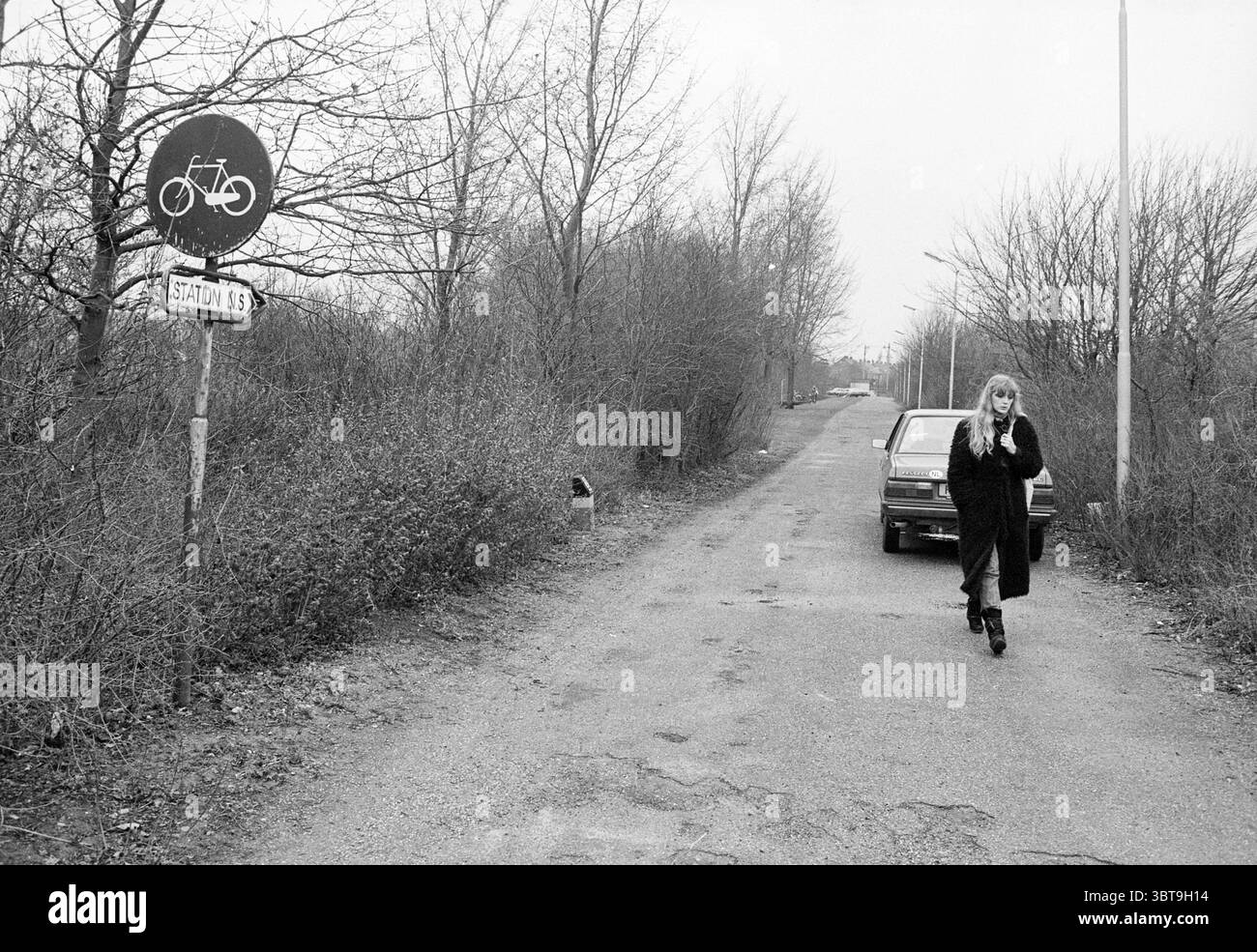 Piste cyclable jusqu'à la station NS Santpoort pistes cyclables cyclistes Santpoort, Whizgle News, Dutch Desk, pays-Bas, 1950 - 2000 on 01-03-1983. Ces rubriques sont illustrées dans l'image. La scène représente une route désolée avec une surface non pavée, flanquée d'arbres clairsemés et d'arbustes épineux. L'atmosphère générale est grise et nuageuse, véhiculant un sentiment de calme et peut-être un soupçon de mélancolie. D'un côté, un panneau de signalisation indiquant une piste cyclable contraste fortement avec la verdure environnante, sa forme circulaire et son symbole blanc sont bien en évidence sur la toile de fond silencieuse. À proximité, an o Banque D'Images