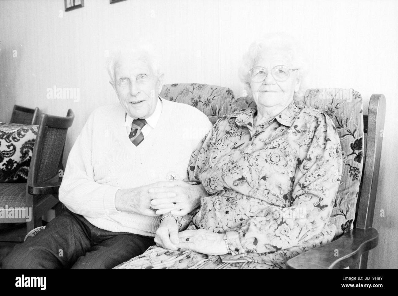 Couple de 65 ans de Essen IJmuiden couples IJmuiden Nederland, Whizgle News, Dutch Desk, The Netherlands, 1950 - 2000 le 19-11-1989. L'image contient ces rubriques. La scène met en scène un couple âgé assis étroitement ensemble sur un canapé à motifs floraux moelleux. L'homme, avec un sourire doux et des cheveux blancs doux, porte un pull tricoté de couleur claire sur une chemise à col, complété par une cravate à motifs. Ses mains sont doucement serrées avec celles de la femme à côté de lui. La femme a les cheveux argentés ondulés et est ornée d'une longue robe florale qui met en valeur des tons doux de rose et de bleu. Elle porte Banque D'Images