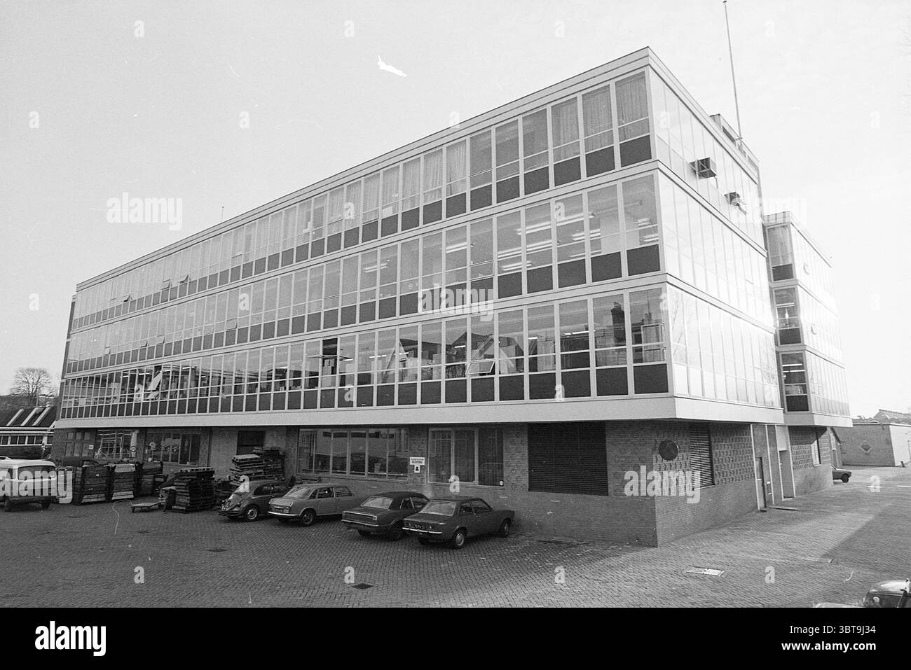 Extérieur EMI Holland Tulpenkade Exterior Haarlem Buildings, Whizgle News, Dutch Desk, pays-Bas, 1950 - 2000 on 13-12-1976. L'image inclut ces rubriques. La scène présente un grand bâtiment moderne caractérisé par son utilisation intensive du verre, permettant un aspect lumineux et ouvert. La structure est principalement composée de plusieurs étages, chacun bordé de grandes fenêtres qui reflètent la lumière et les environs, suggérant un style architectural contemporain. L'extérieur présente une palette monochromatique de gris et de blanc, mettant l'accent sur une esthétique industrielle épurée. Au premier plan, par Banque D'Images