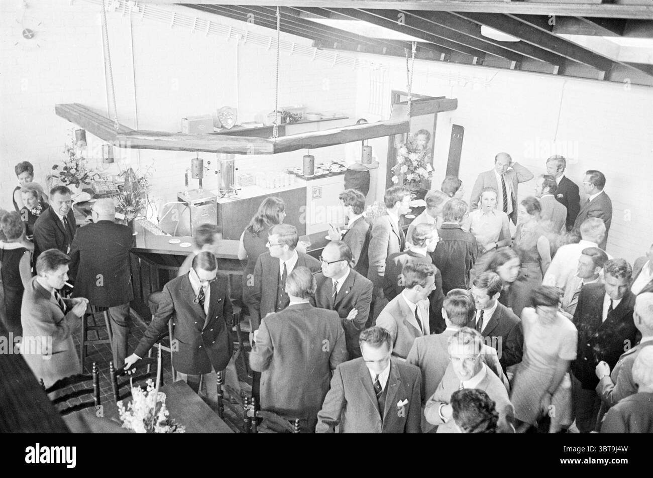 Réception ADO'20 ADO Football, Whizgle News, Dutch Desk, pays-Bas, 1950 - 2000 le 16-05-1970. Voici les rubriques de l'image. La scène est animée d'activités, capturant un rassemblement social animé. Un groupe diversifié de personnes, habillées dans un mélange de vêtements formels et semi-formels, se déplace à travers un espace intérieur rustique. Les couleurs prédominantes sont atténuées, avec des nuances de gris et de noir mêlées à des éclairs occasionnels de couleur provenant des vêtements et accessoires. En arrière-plan, une barre s'étend le long d'un côté, avec des éléments en bois combinés avec des accents métalliques. La verrerie est proprement arr Banque D'Images