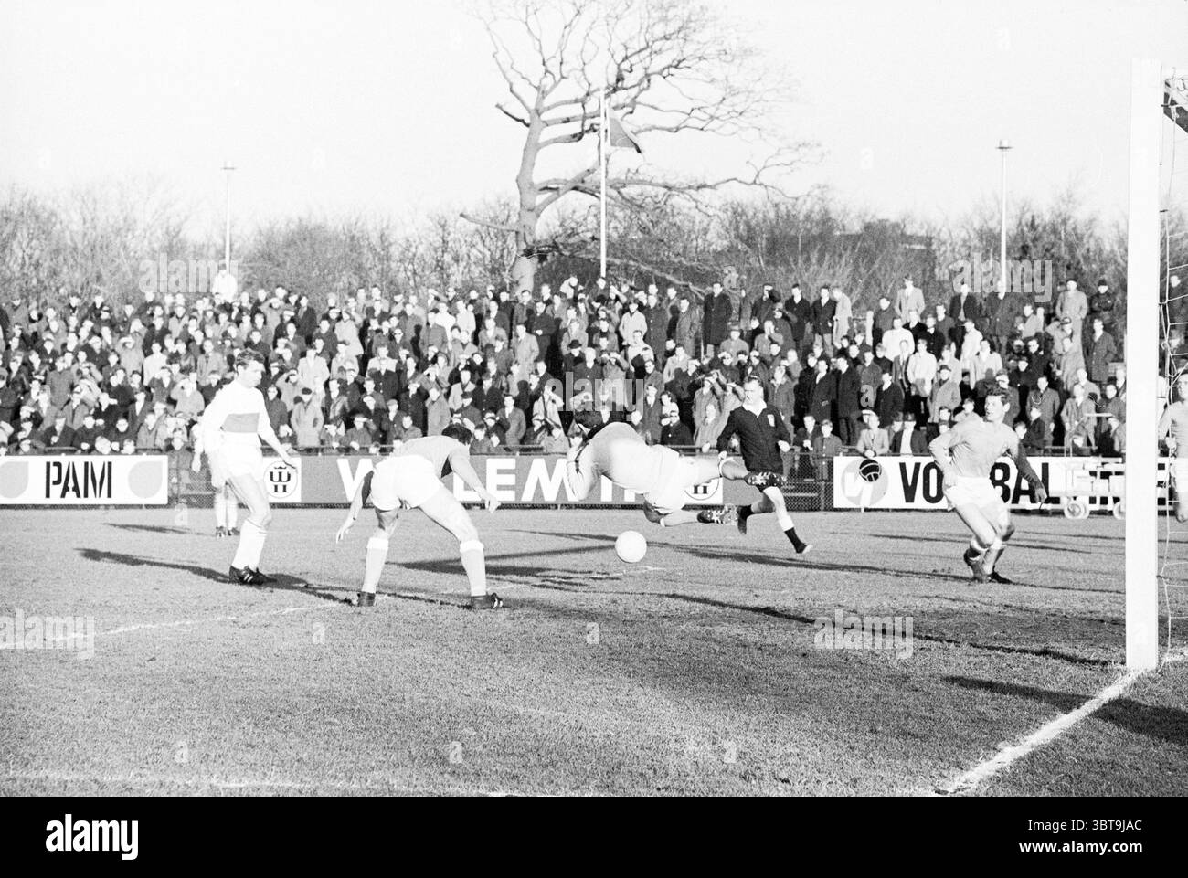 D.E.M. - V.V.B. Football, Whizgle News, Dutch Desk, pays-Bas, 1950 - 2000 le 28-01-1968. Voici les rubriques de l'image. La scène représente un moment intense lors d'un match de football qui se déroule à l'extérieur. Au premier plan, un joueur portant un maillot blanc fait un mouvement pivot, prêt à frapper la balle qui se trouve à quelques mètres. Son corps est légèrement penché vers l'avant, indiquant un éclat d'énergie et de concentration. À sa droite, un autre joueur en blanc semble être en position défensive, tandis que le gardien de but, vêtu d'un uniforme noir, saute pour intercepter le ballon, présentant un affichage dynamique Banque D'Images
