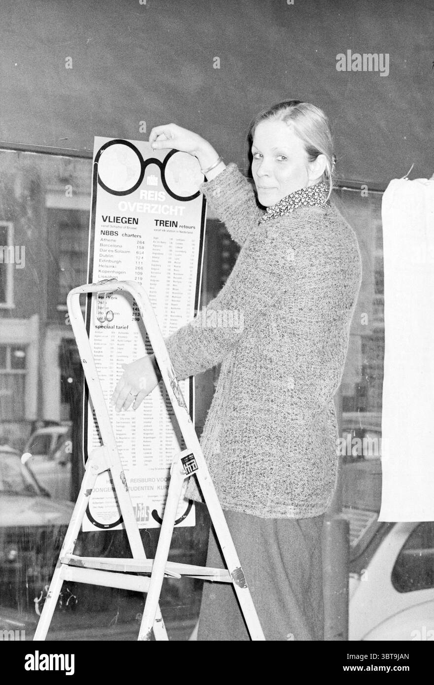 Bureau NBBS Interior Haarlem Botermarkt pays-Bas, Whizgle News, Dutch Desk, pays-Bas, 1950 - 2000 le 13-02-1976. L'image montre ces rubriques. Une femme se tient sur un escabeau, engagée dans l'acte d'accrocher un grand signe. Elle a les cheveux longs et porte un pull gris texturé douillet associé à un pantalon sombre et ample. L'expression sur son visage est une expression de concentration, légèrement souriante lorsqu'elle se penche vers l'avant pour positionner le signe avec précision. Le cadre semble être un environnement urbain, peut-être près d'un magasin ou d'un café, comme le suggère la grande fenêtre en arrière-plan. Dehors, flou Banque D'Images