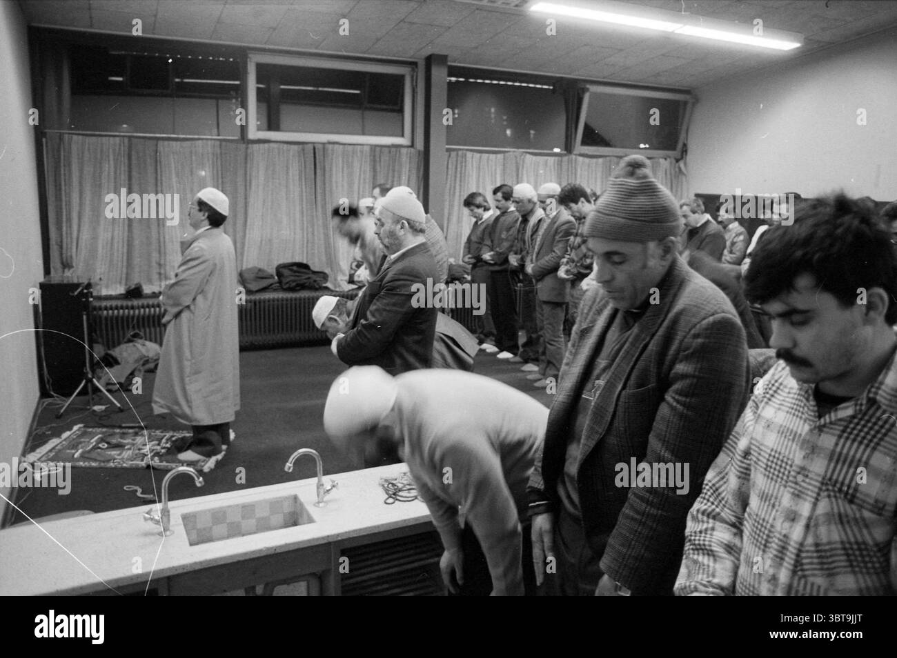 Prière à la mosquée H'lem Haarlem pays-Bas, Whizgle News, Dutch Desk, pays-Bas, 1950 - 2000 le 10-03-1992. L'image contient ces rubriques. La scène représente un rassemblement solennel dans un espace intérieur, probablement un centre communautaire ou un lieu désigné pour le culte. L'éclairage est coupé, provenant de luminaires suspendus qui éclairent la pièce sans être trop lumineux, projetant des ombres douces sur le sol. Au premier plan, un petit groupe d'individus est vu agenouillé dans la prière, leur tête inclinée dans la vénération. Ils portent un mélange de vêtements traditionnels, y compris des châles et des chapeaux, qui Banque D'Images