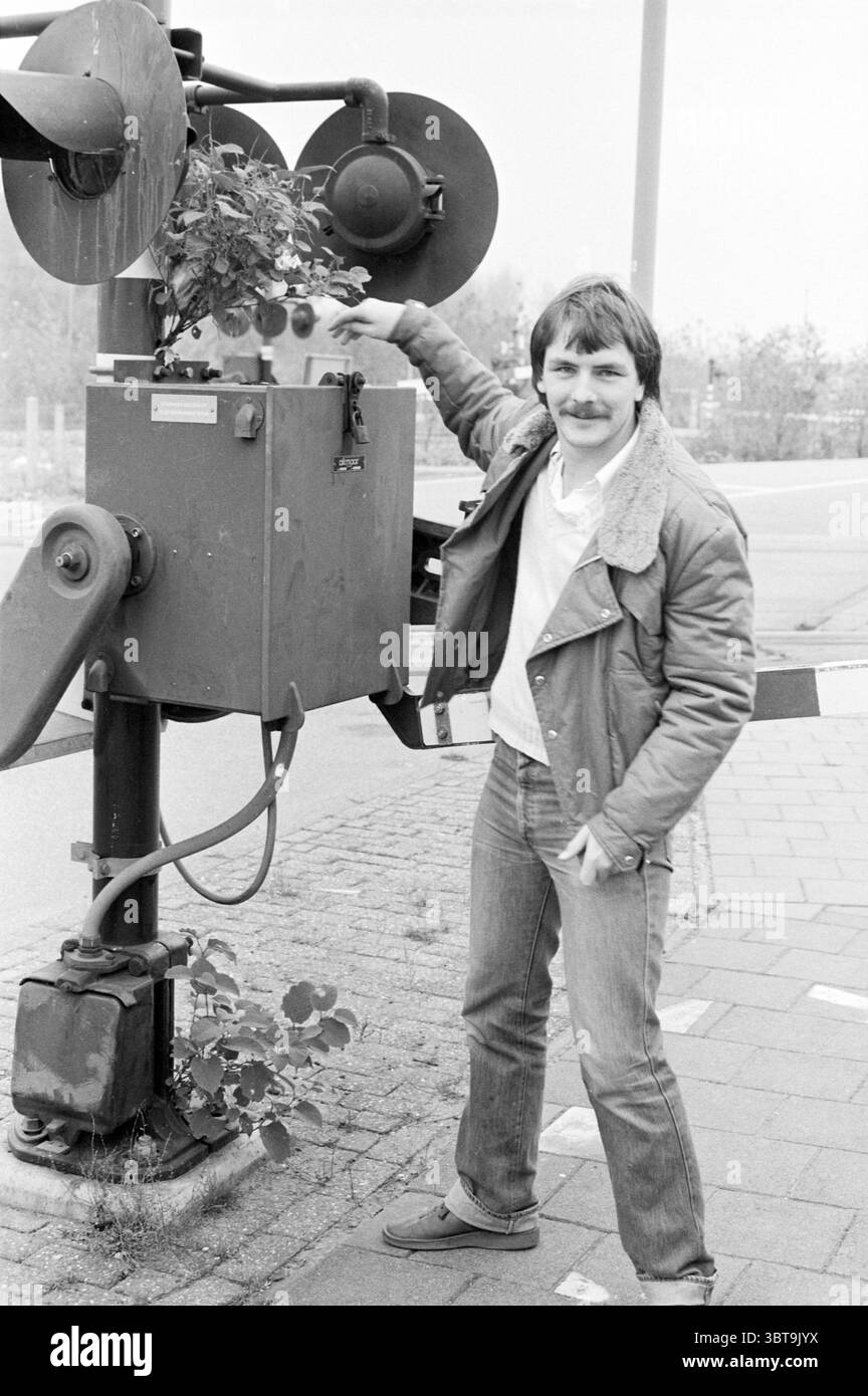 Arbre poussant dans les chemins de fer à lumière clignotante, Whizgle News, Dutch Desk, The Netherlands, 1950 - 2000 le 04-11-1982. Ces rubriques apparaissent dans l'image. Dans une scène monochrome silencieuse, un homme se tient confiant à côté d'un vieux dispositif de signalisation, probablement lié à des passages à niveau. L'homme a une moustache soigneusement soignée et arbore une tenue décontractée qui comprend une chemise de couleur claire sous une veste floue, jumelée à un Jean ajusté. Sa posture est détendue alors qu'il s'appuie de manière ludique contre l'appareil, une main reposant dessus tandis que l'autre est sur sa hanche. Le dispositif de signalisation présente une structure boxy avec se Banque D'Images
