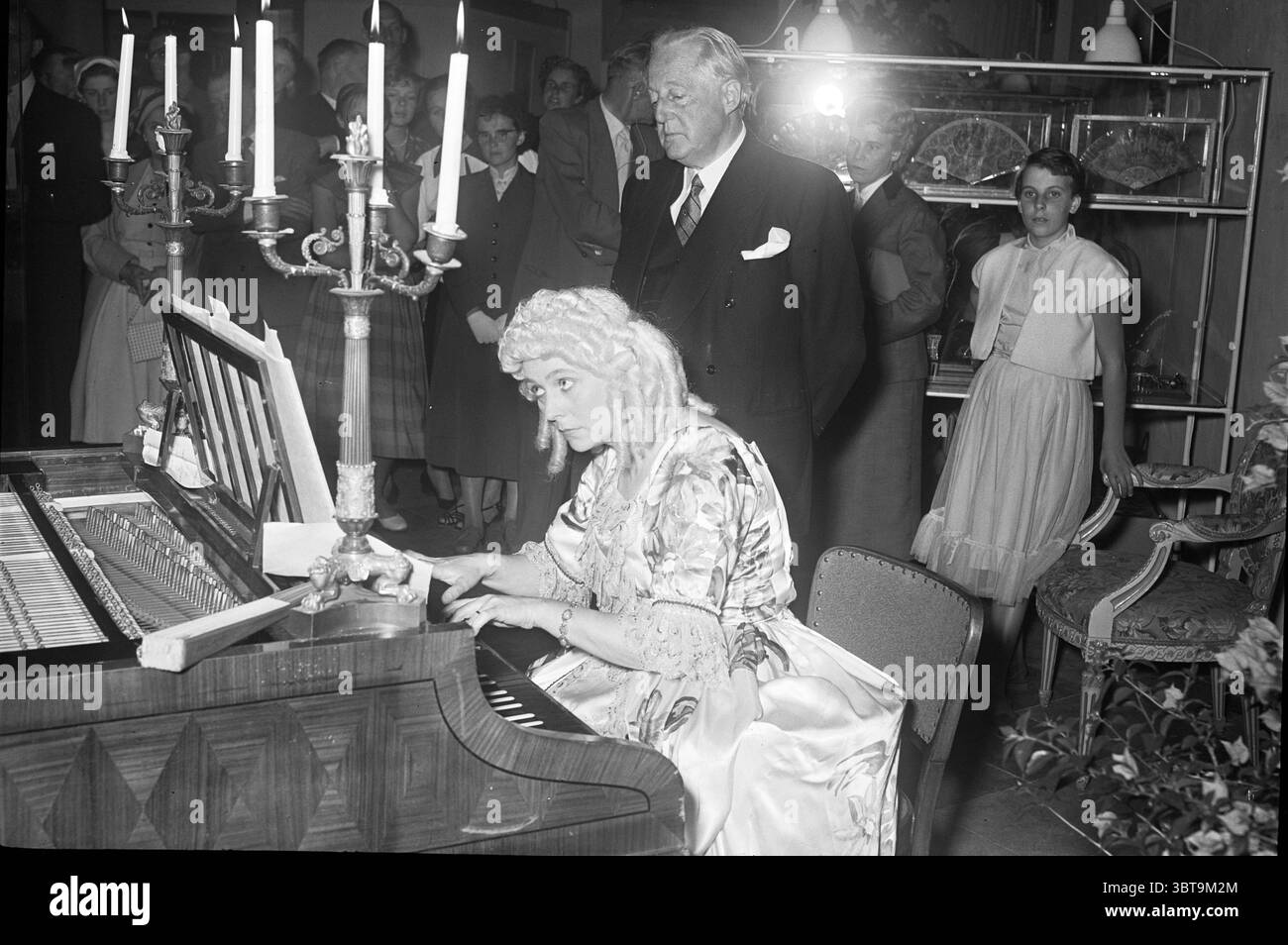 Récital de piano., Whizgle News, Dutch Desk, pays-Bas, 1950 - 2000. L'image inclut ces rubriques. La scène capture un rassemblement vintage dans une pièce bien décorée qui respire l'élégance et un air subtil de nostalgie. À l'avant-garde, une femme s'assoit gracieusement devant un piano à queue, sa robe longue et fluide chatoyante avec des motifs complexes et des teintes douces qui suggèrent à la fois raffinement et drame. Ses cheveux sont coiffés dans des boucles douces, peut-être ornés d'une pièce décorative qui complète sa tenue vestimentaire, attirant l'attention sur son expression focalisée pendant qu'elle joue. Autour d'elle se trouvent plusieurs individus, certains Banque D'Images