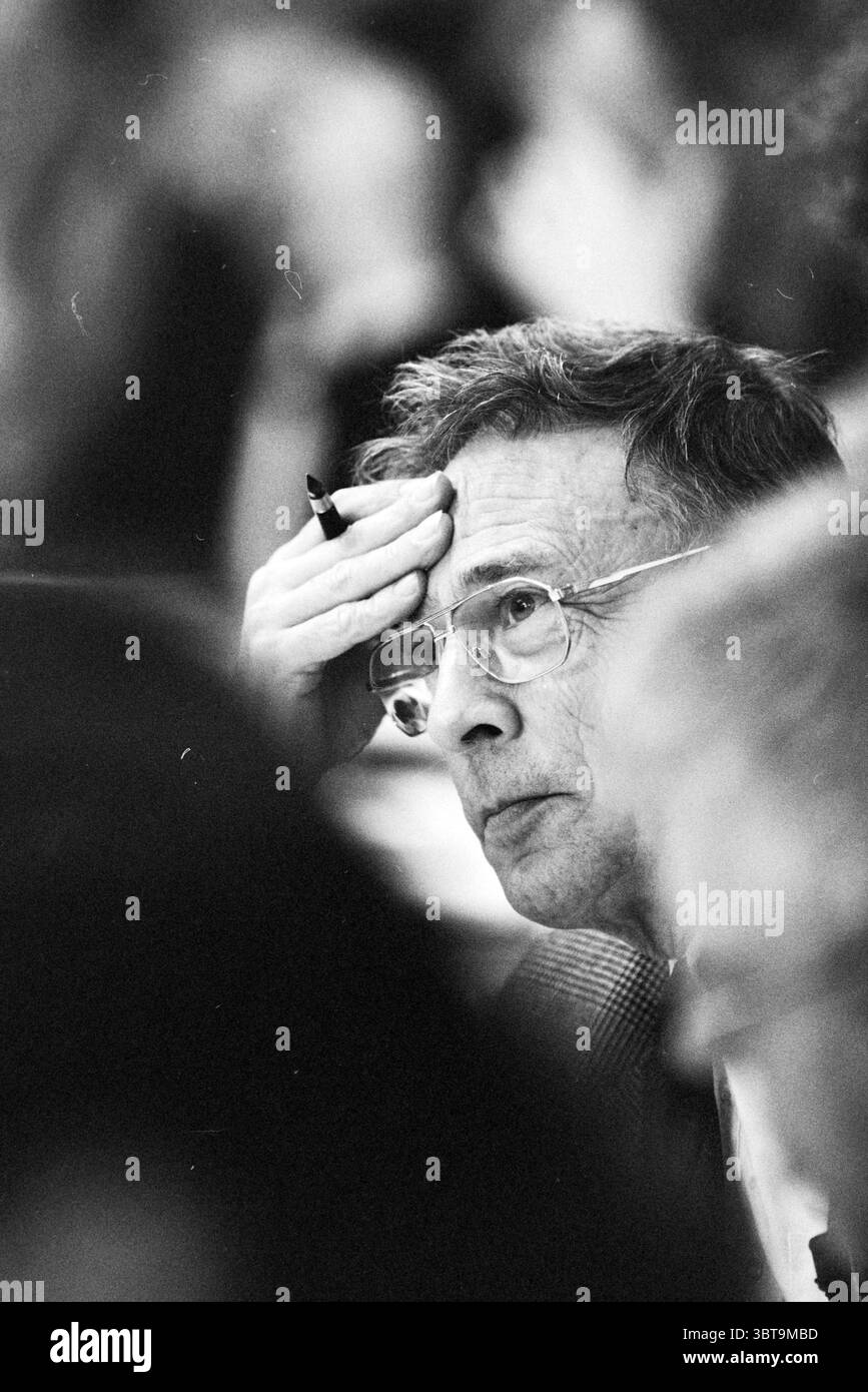 conseil du budget H'lem Haarlem pays-Bas, Whizgle News, Dutch Desk, pays-Bas, 1950 - 2000 le 22-11-1994. Voici les rubriques de l'image. Dans un cadre faiblement éclairé, la scène capture un monsieur plus âgé assis parmi un groupe d'individus flous. Son expression révèle une humeur contemplative ; une main repose soigneusement sur son front tandis que l'autre tient un stylo près de ses lèvres. Ses lunettes aperçoivent la lumière environnante, soulignant son regard perspicace alors qu'il regarde, profondément dans la pensée. L'arrière-plan est rempli de formes douces et diffuses qui suggèrent la présence de Banque D'Images