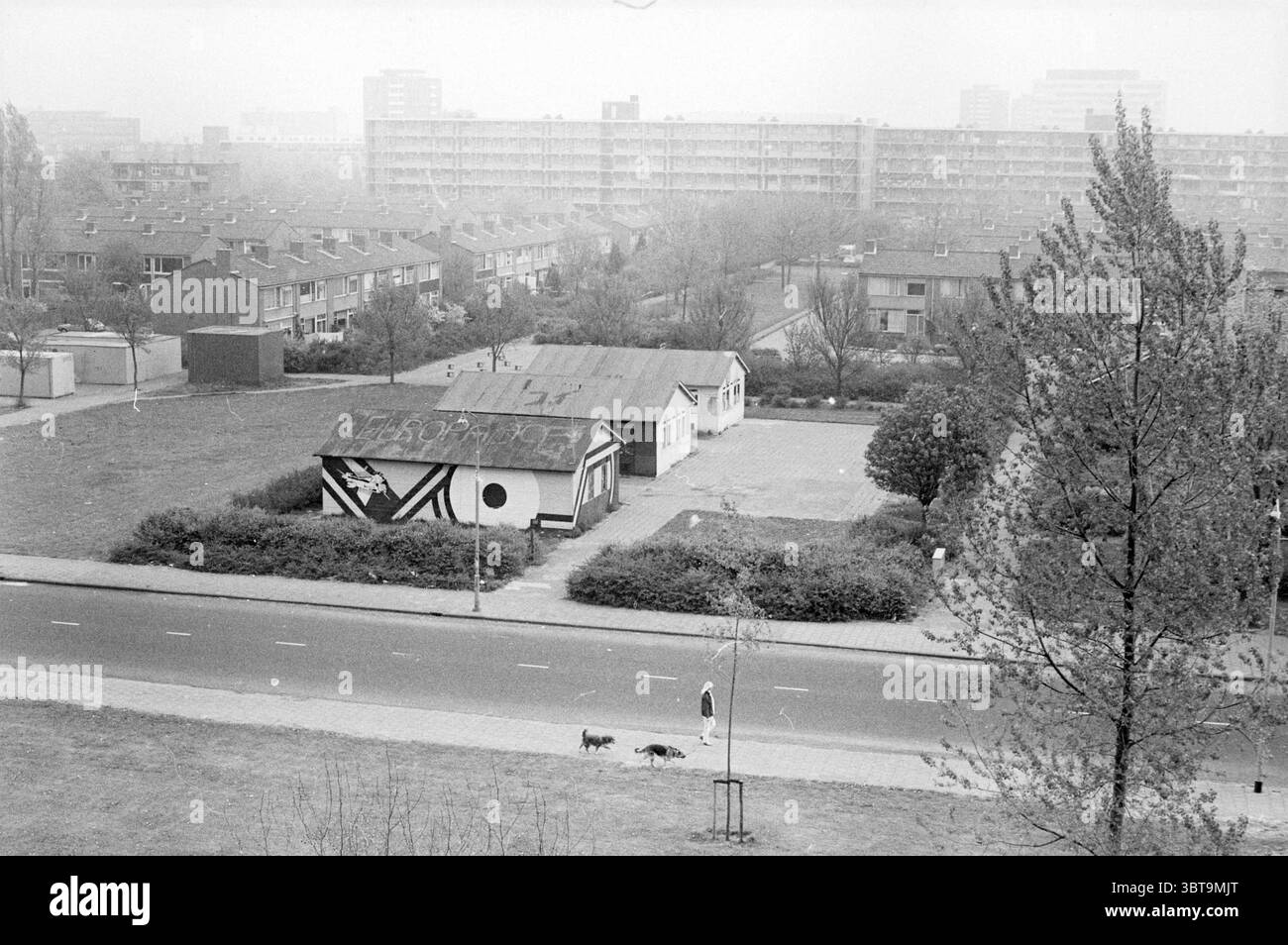 Bases jeunesse Schalkwijk Jeunesse et jeux jeunesse, Whizgle News, Dutch Desk, pays-Bas, 1950 - 2000 le 04-05-1984. Ces rubriques apparaissent dans l'image. La scène présente un paysage urbain caractérisé par un sentiment de calme et d'immobilité. Au premier plan, un bâtiment modeste se distingue par ses motifs géométriques noirs et blancs distinctifs d'un côté, contrastant avec son environnement plus discret. Son toit semble légèrement altéré, suggérant à l'âge et peut-être à la négligence. Autour du bâtiment, une étendue d'herbe s'étend, ponctuée par des taches de verdure sauvage et quelques arbres th Banque D'Images